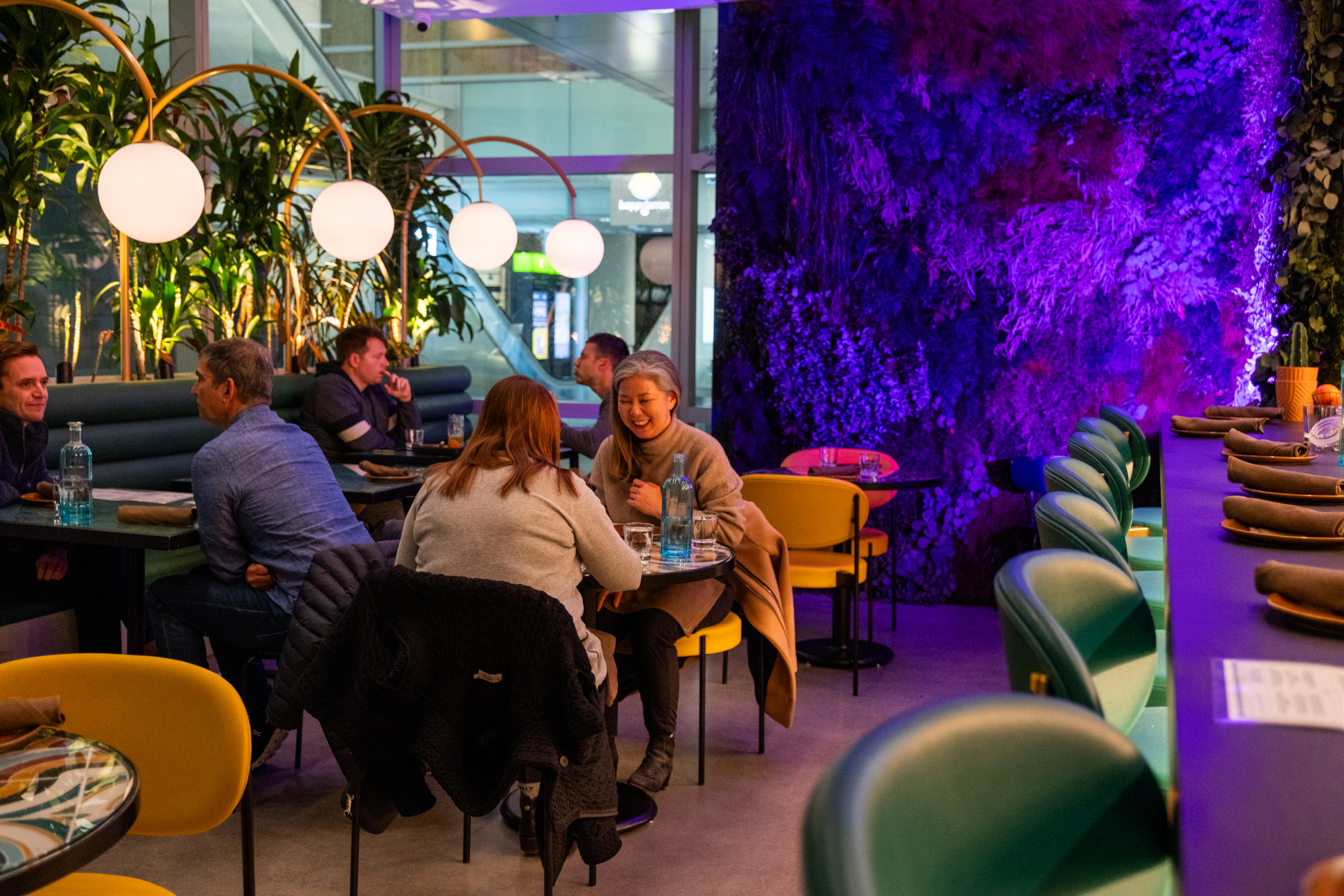 A cozy restaurant scene with people sitting at tables, surrounded by plants and warm lighting. The decor includes green chairs and a wall with vibrant purple lighting.