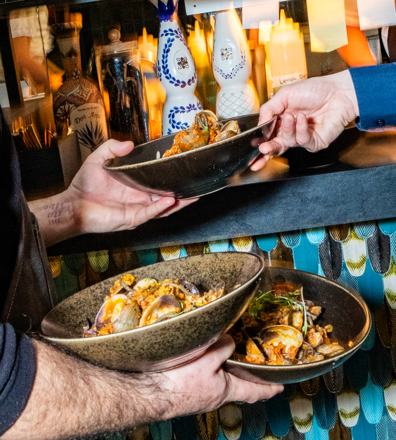 Two hands exchange bowls filled with clams and garnished food. Bottles and condiments are in the background, suggesting a vibrant restaurant setting.