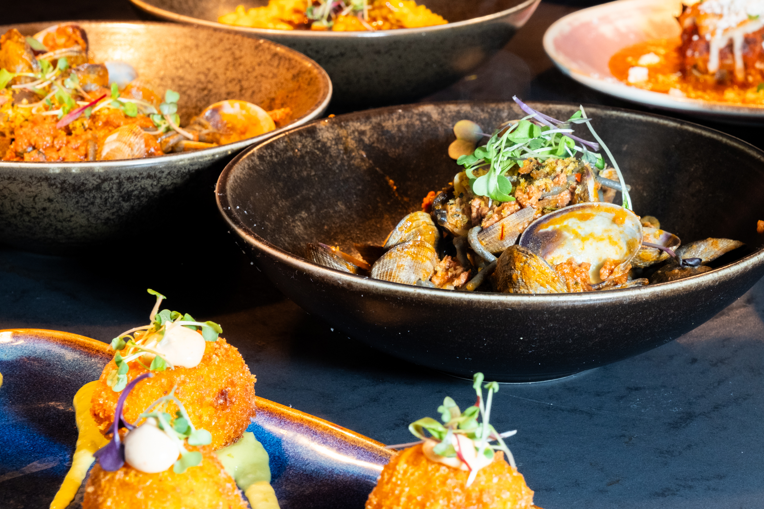 The image shows elegant dishes featuring clams with herbs in black bowls and breaded appetizers topped with garnishes on a dark plate.