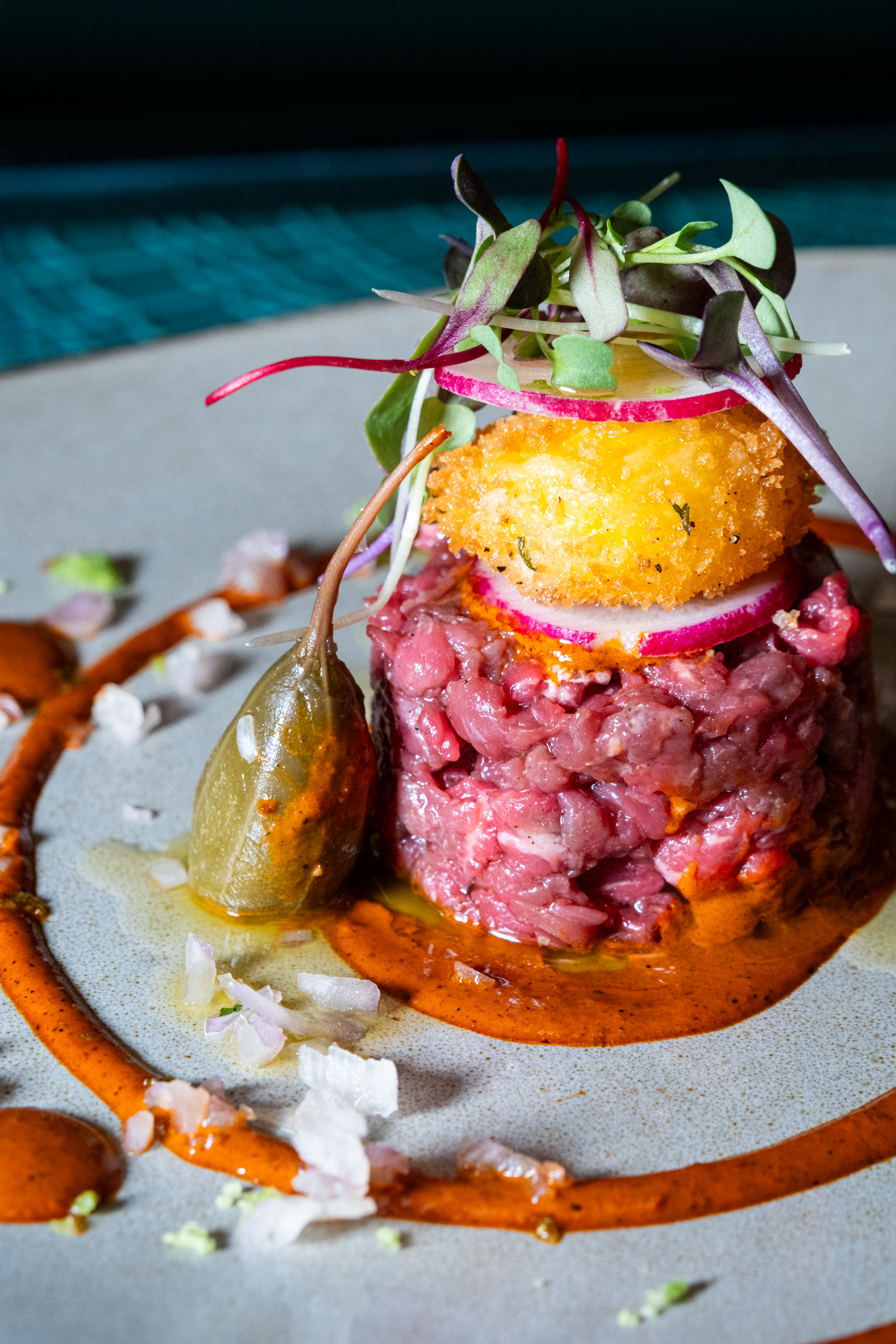 A gourmet dish featuring a stack of raw minced meat topped with a fried ball, radish slices, and microgreens, surrounded by sauce and garnished with a caperberry.