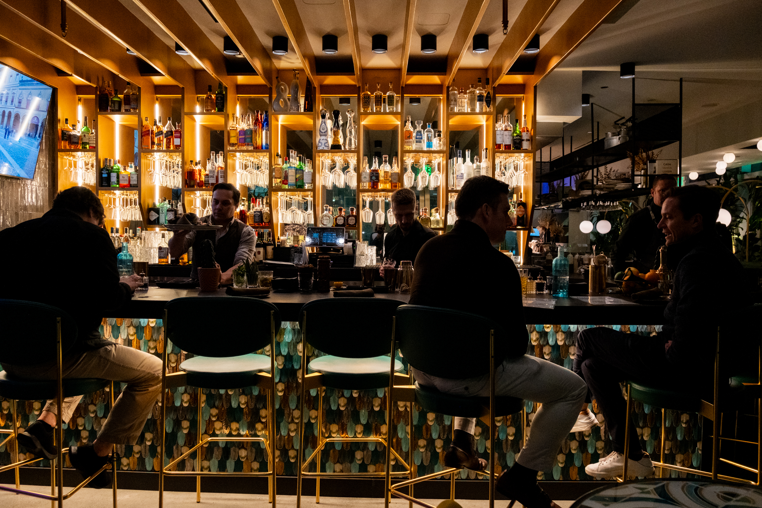 A lively bar with glowing shelves of liquor in the background, where patrons sit on high stools. The ambiance is warm, with a bartender serving drinks.