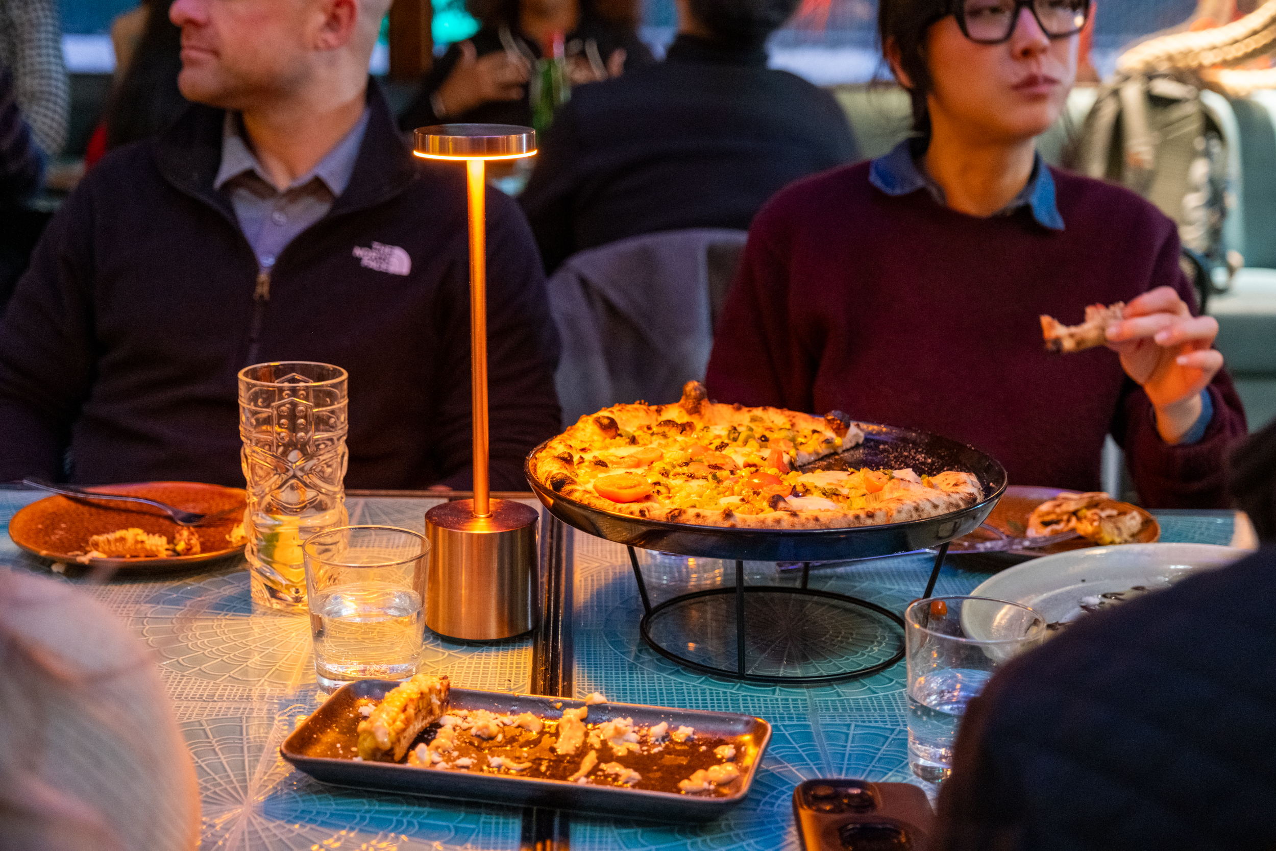 Two people sit at a table with pizza, a glowing table lamp, and drinks. One person is eating a slice, while dishes and an empty tray are visible.