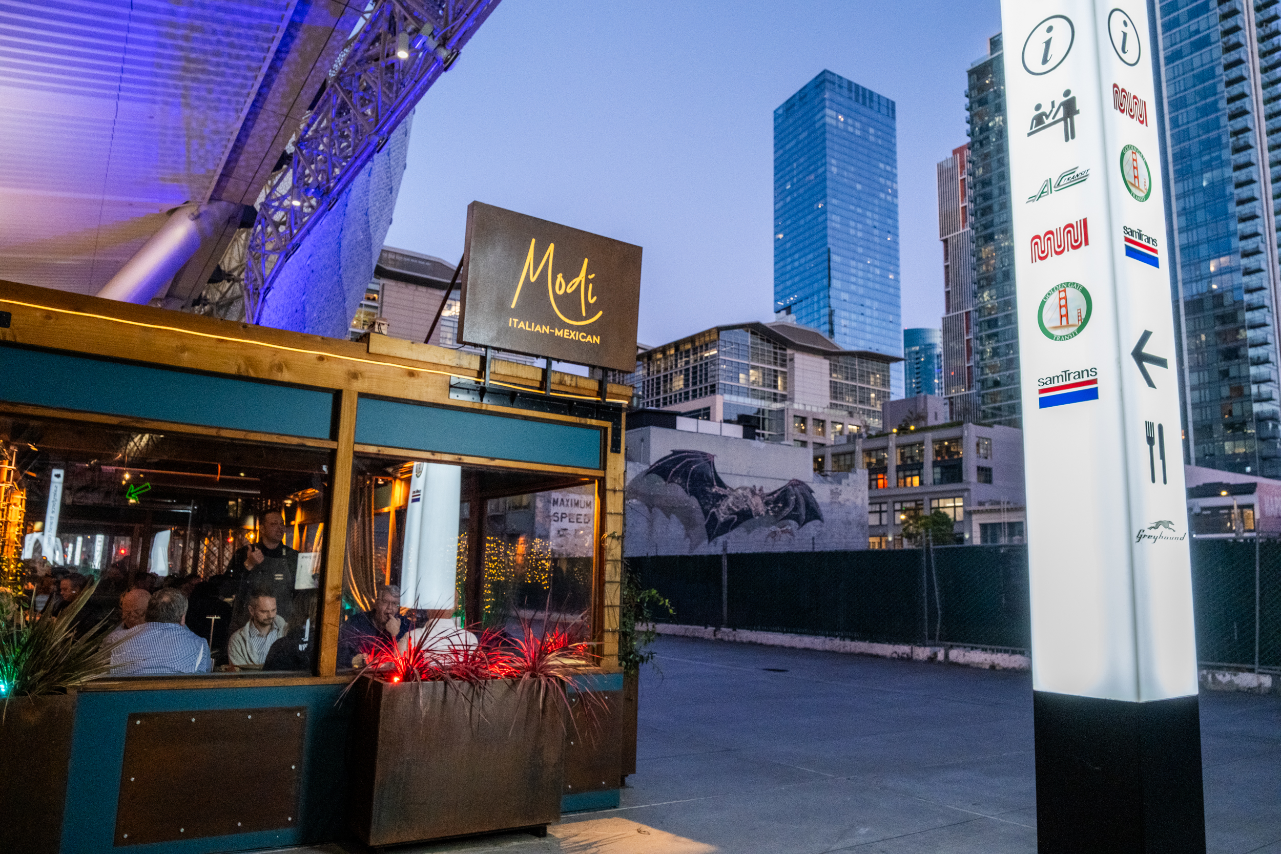 A modern street scene features a restaurant named Modi, an outdoor dining area, and a tall sign with transit icons. Skyscrapers tower in the background under an evening sky.