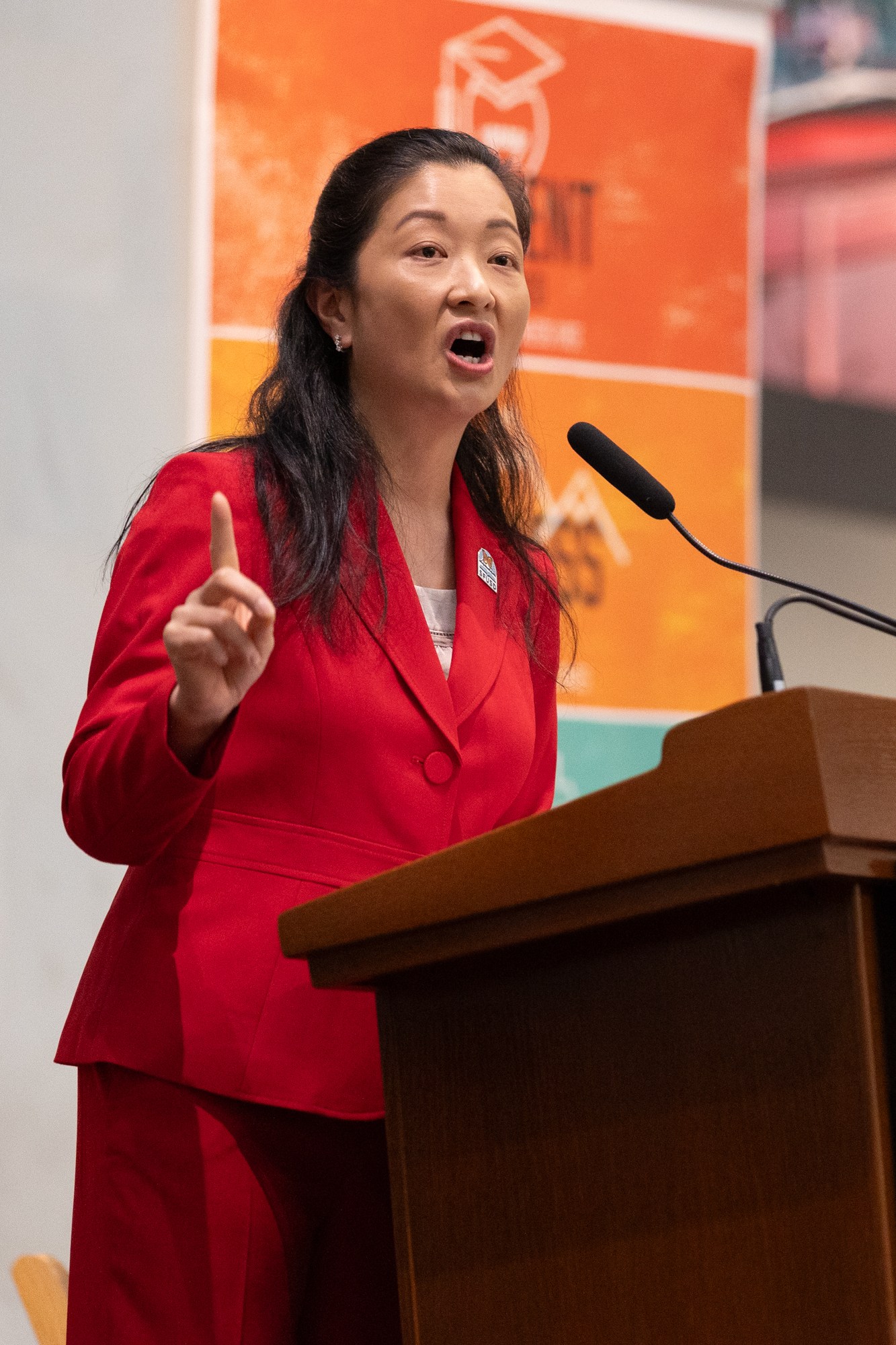 A woman in a red suit speaks passionately at a podium with a seal. A flag is partially visible behind her, and another person is seated in the background.