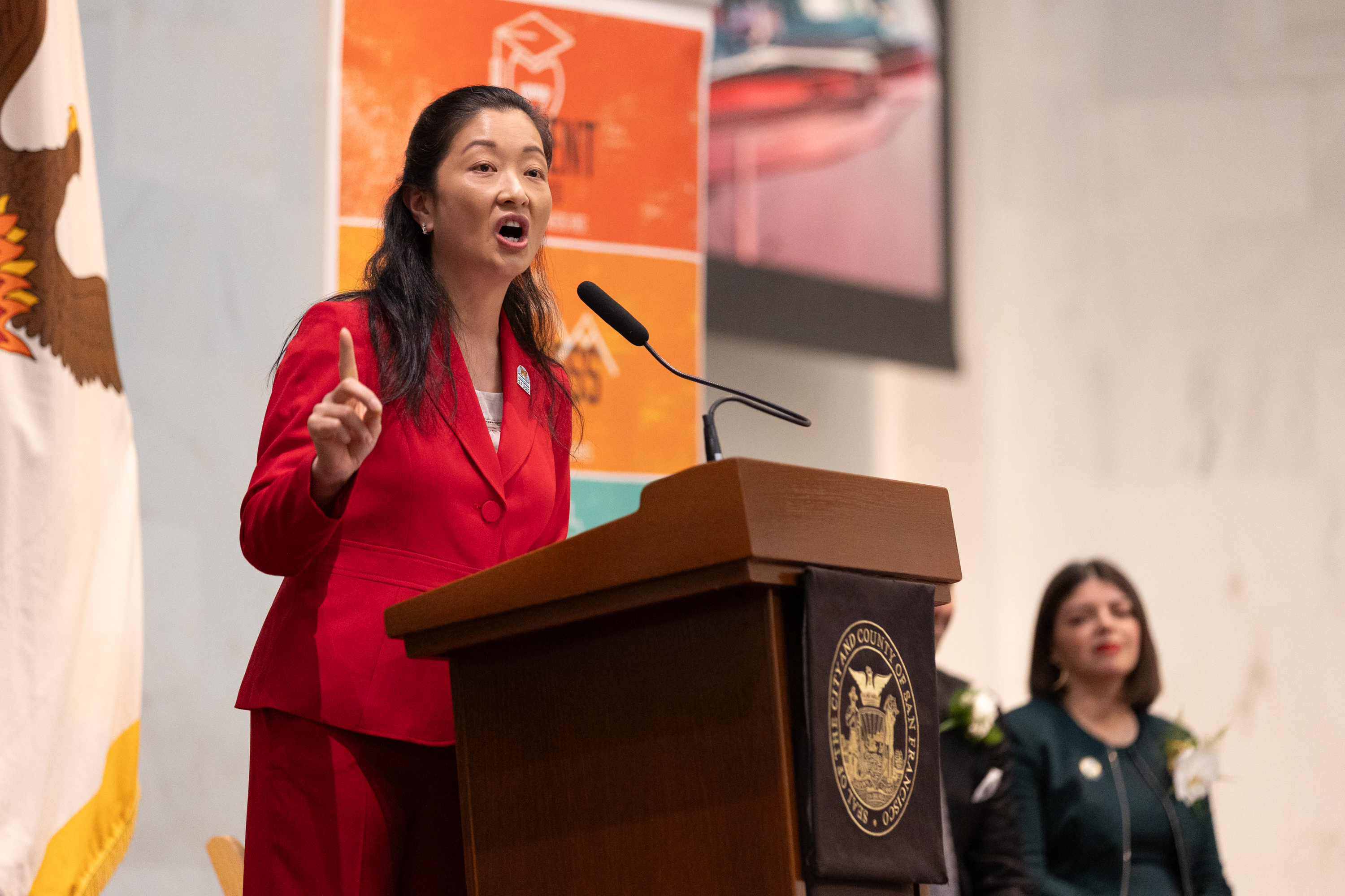 A woman in a red suit speaks passionately at a podium with a seal. A flag is partially visible behind her, and another person is seated in the background.