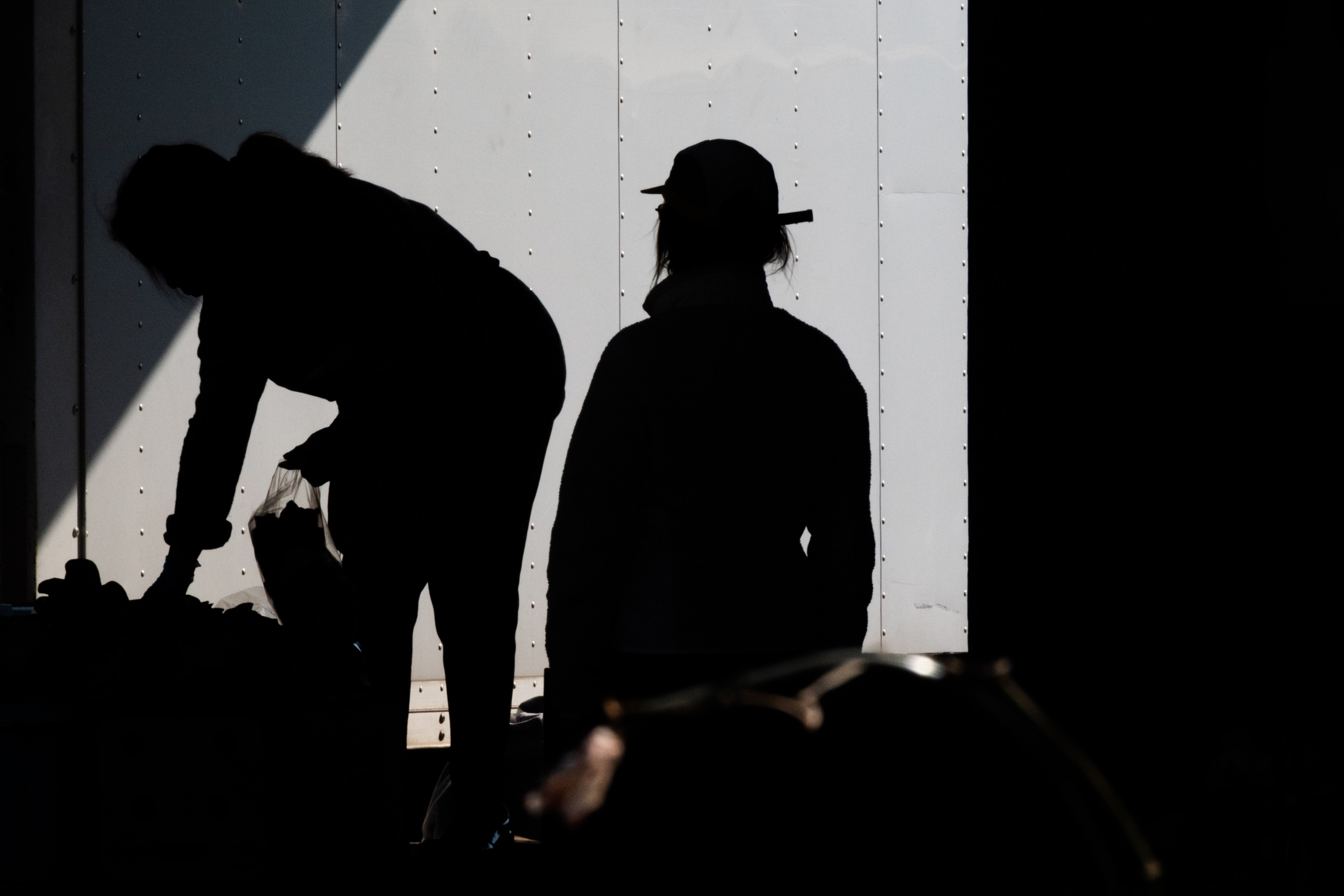 The image shows the silhouettes of two people against a bright, vertical background. One person appears to be bending over, while the other stands upright wearing a hat.