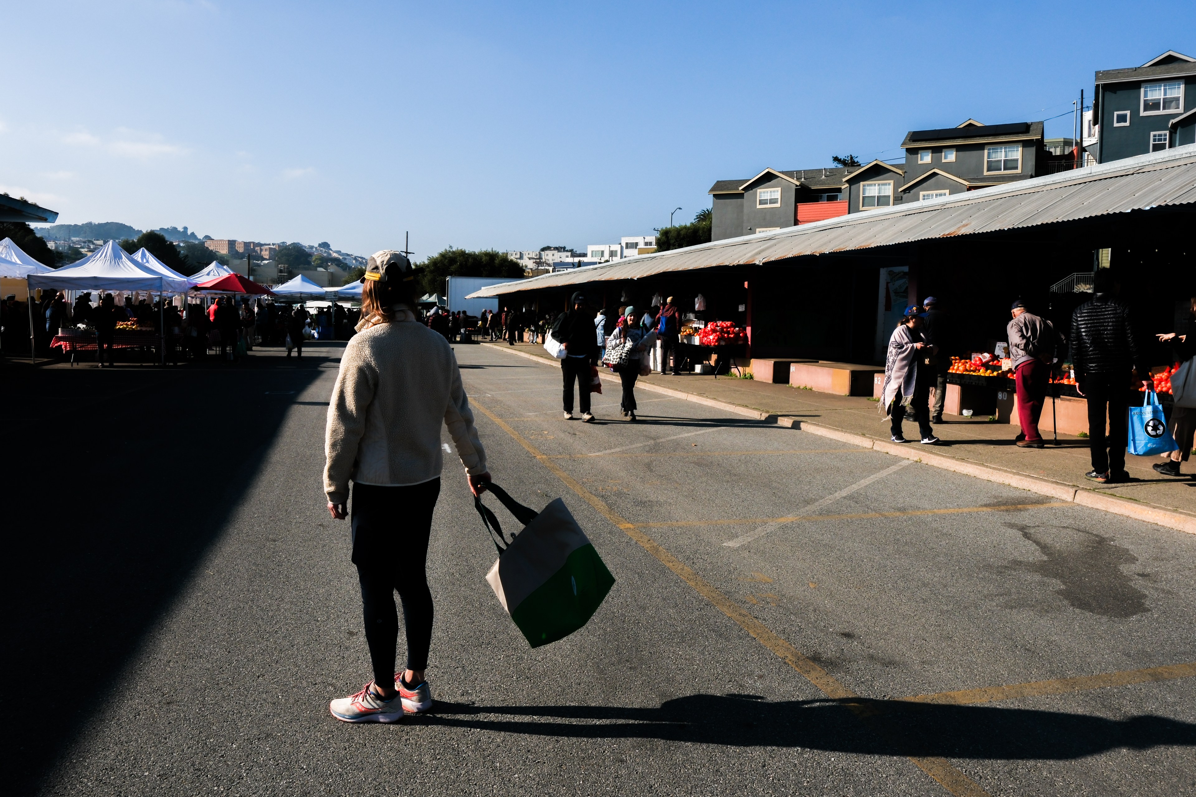 A market scene shows people walking among stalls with white tents, under a clear blue sky. A person in the foreground holds a shopping bag.