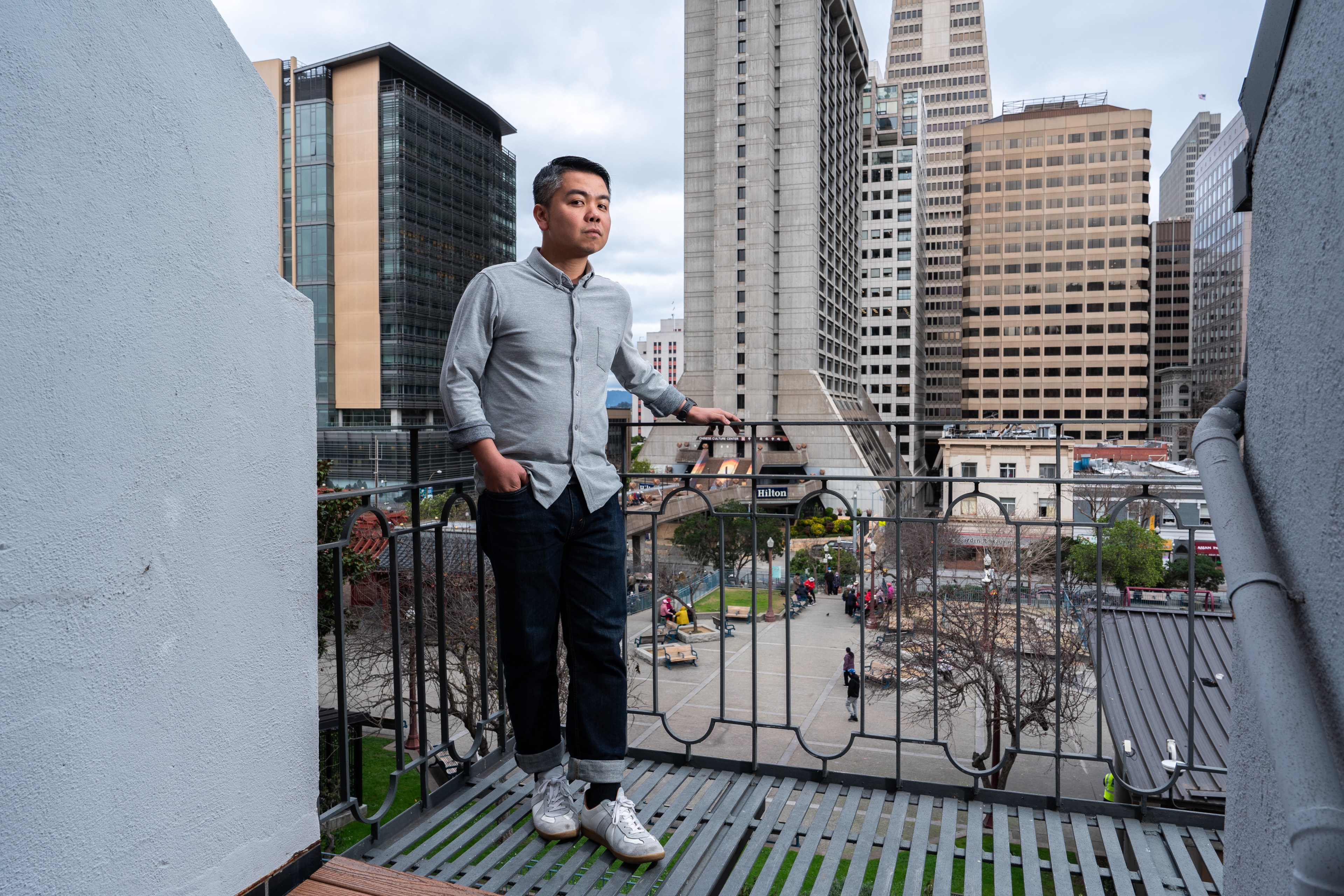 A person in a gray shirt and jeans stands on a balcony overlooking a cityscape with tall buildings and a small park below. The sky is overcast.