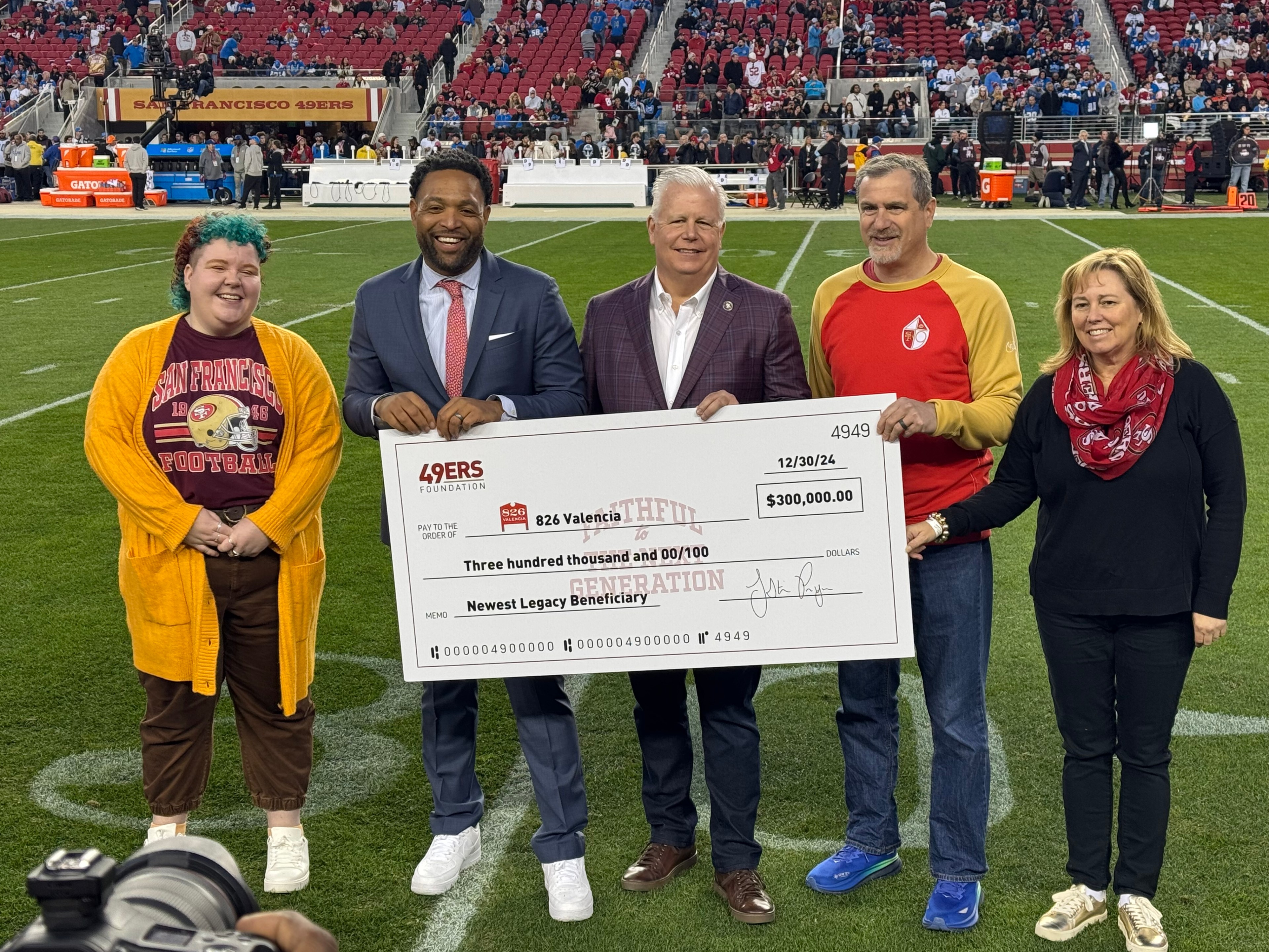 Five people stand on a football field, smiling and holding an oversized check for $300,000 from the 49ers Foundation, in front of a crowd in the stadium.