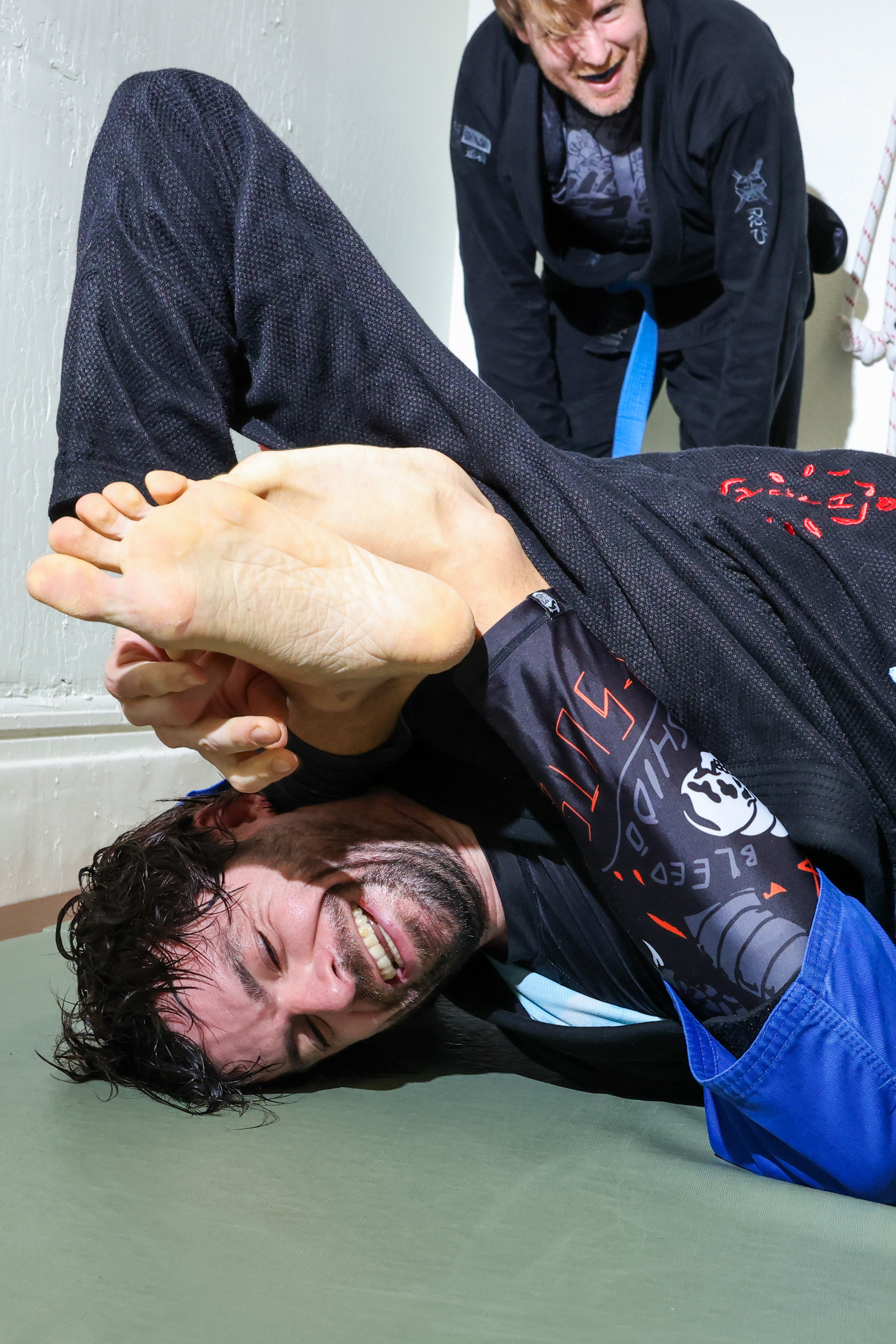 Two people in martial arts uniforms are practicing jiu-jitsu; one is smiling playfully while attempting a complicated leg maneuver on the mat.