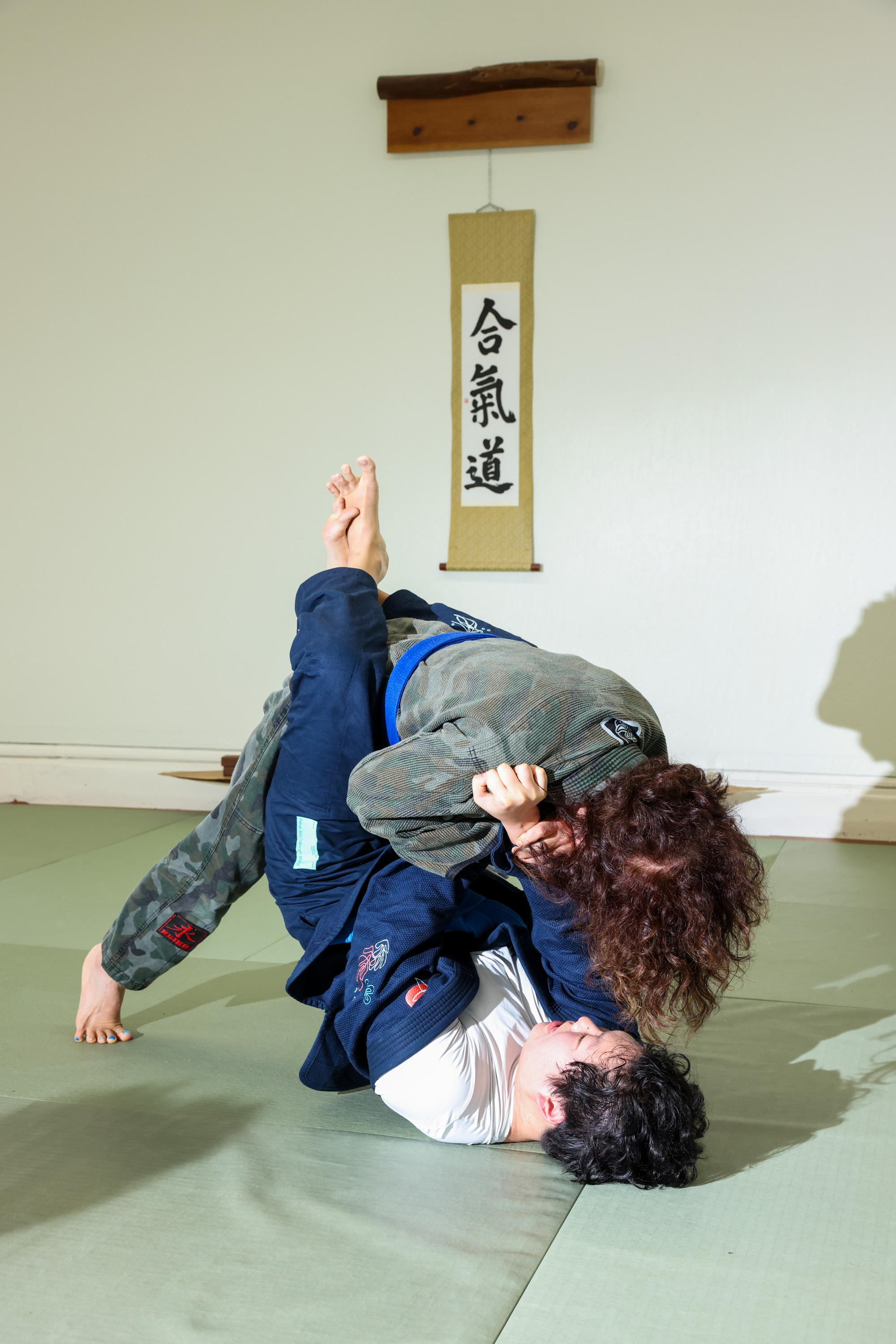 Two people practice martial arts on a mat, one in a blue gi and the other in green camo. They are grappling, with a banner displaying Japanese characters on the wall.