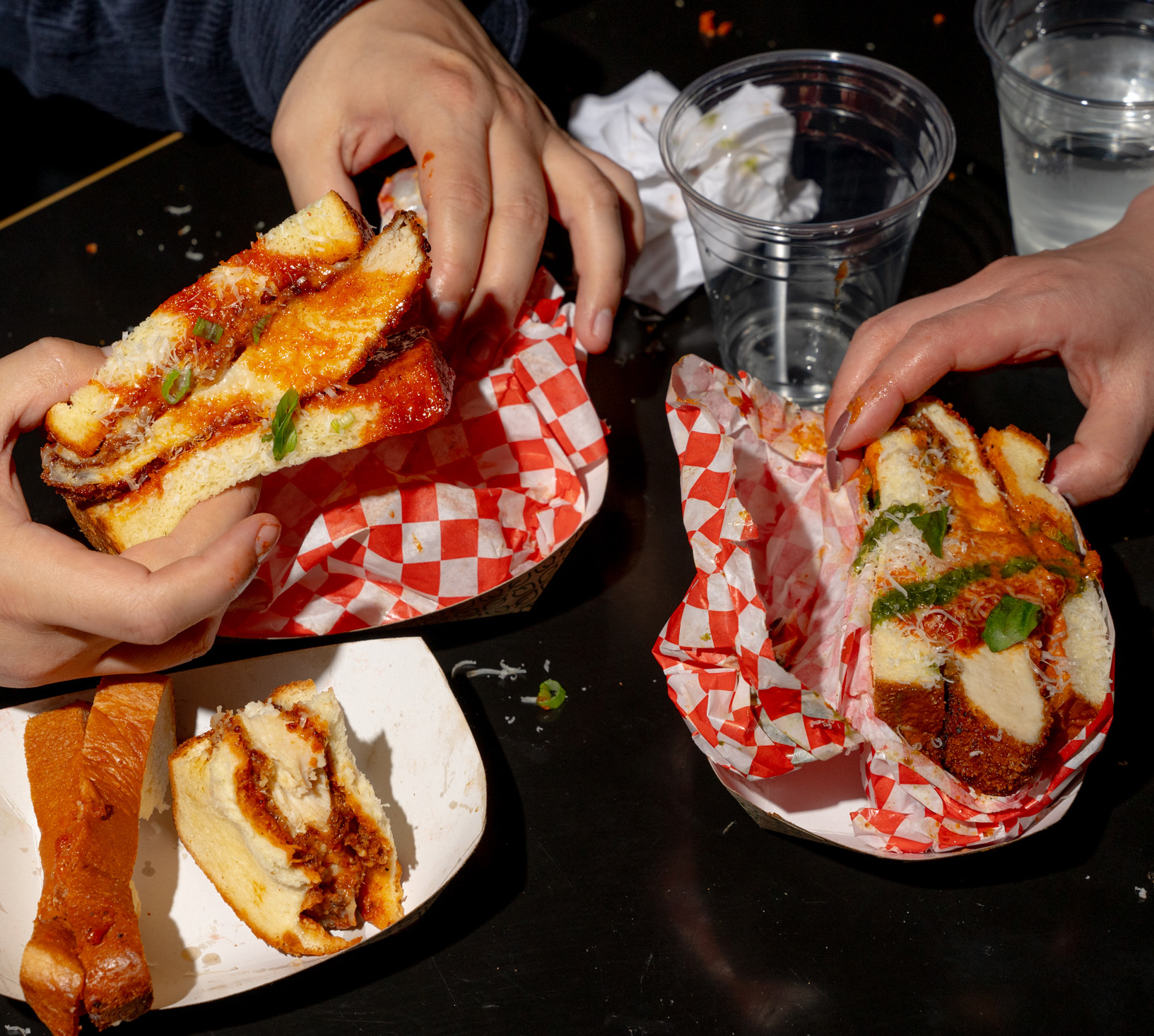 Two people hold sandwiches filled with crispy chicken, cheese, and basil. The sandwiches are wrapped in red-checkered paper with drinks nearby.