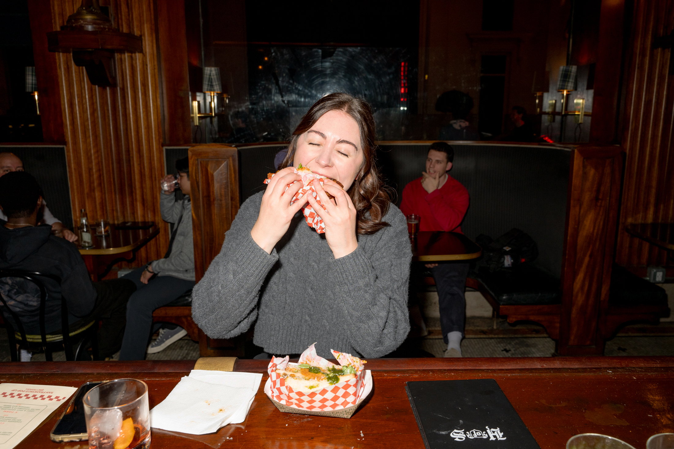 A woman in a gray sweater eagerly bites into a sandwich at a bar with wooden interiors. Others are seated in the background, and a drink is on the counter.