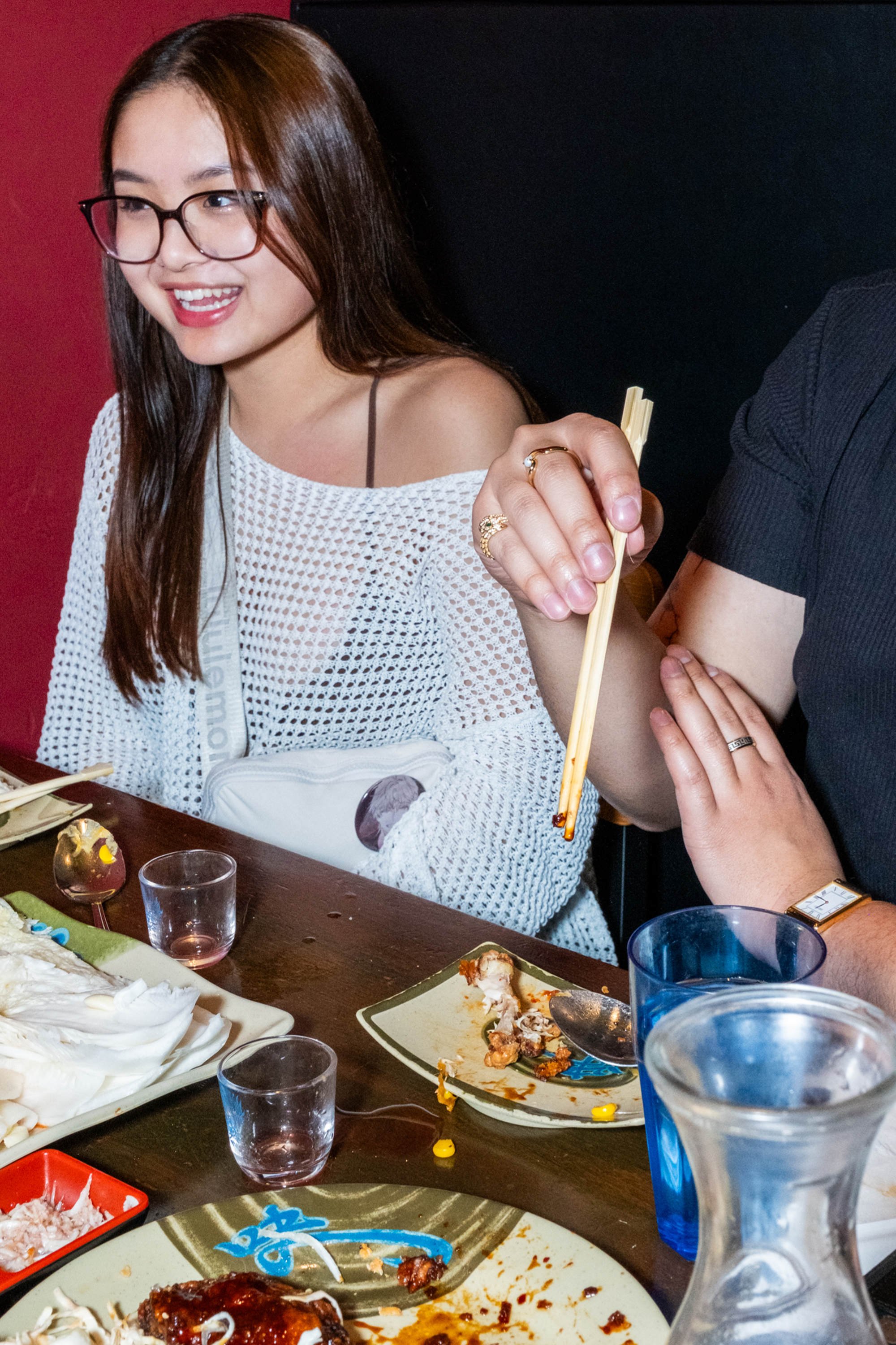 A woman in glasses smiles at a dining table with empty plates and drinks. A hand beside her holds chopsticks, and there's a carafe of water nearby.