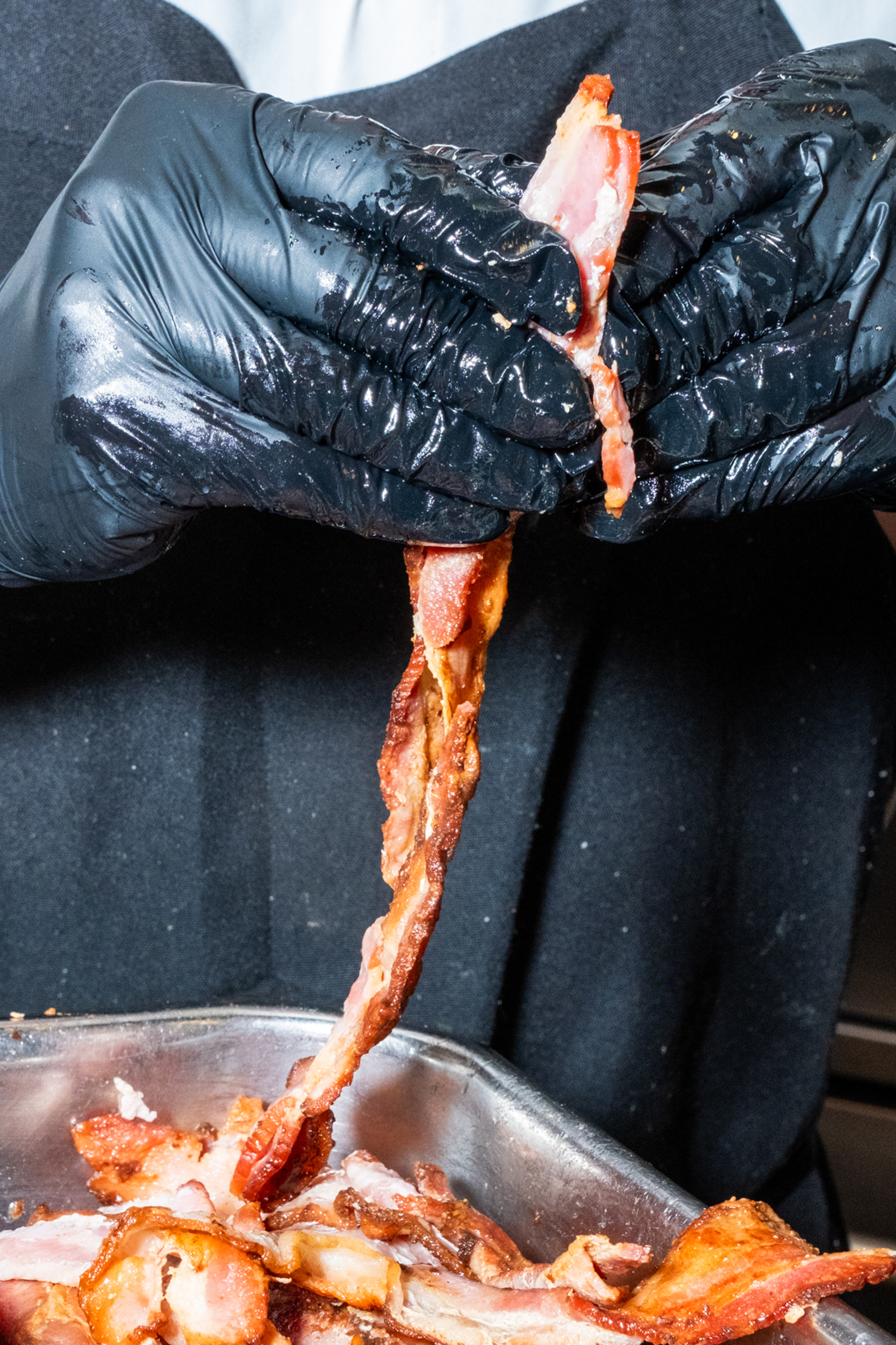 A person wearing black gloves is holding and pulling apart strips of cooked bacon over a metal tray filled with more bacon.