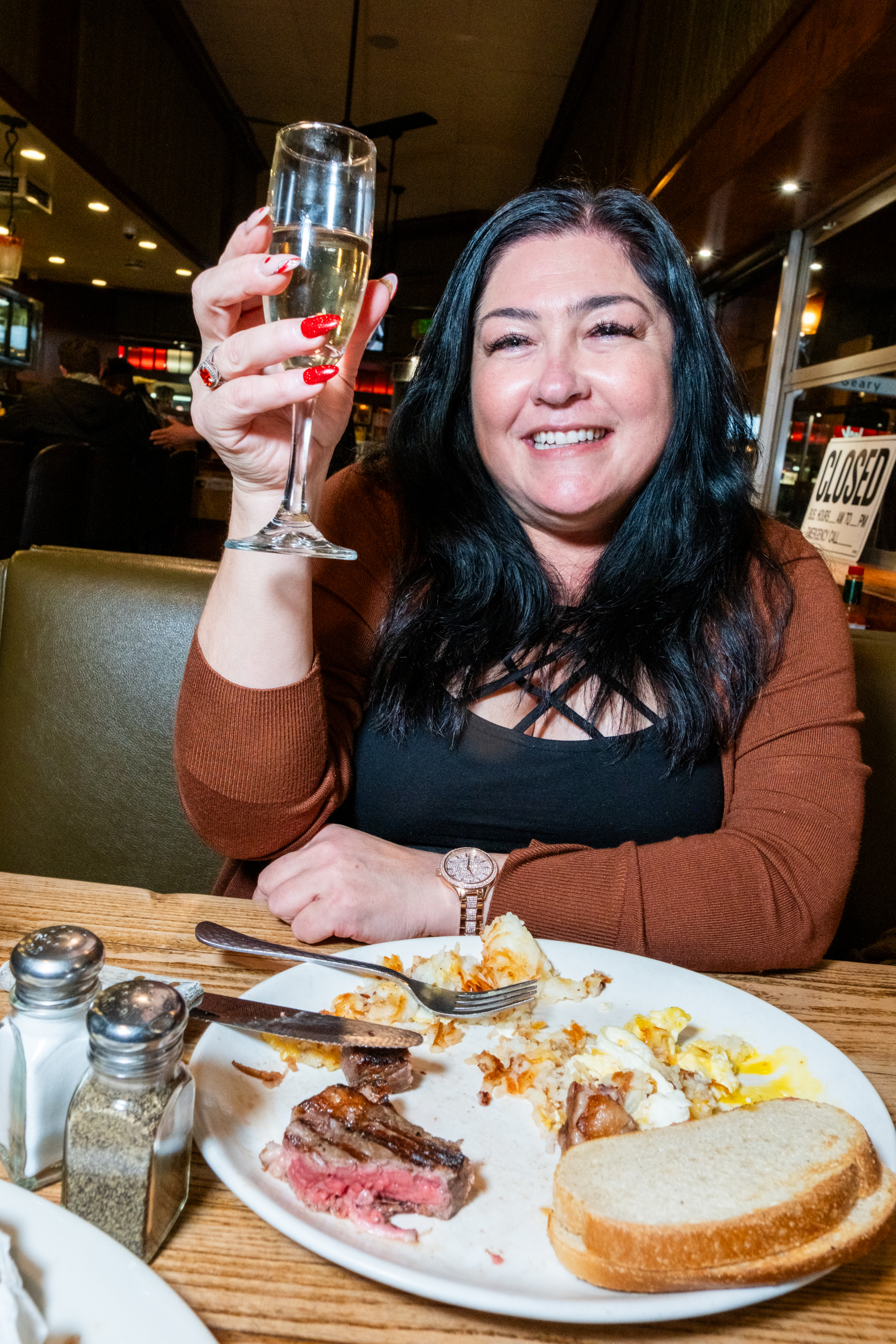 A woman with long dark hair smiles, raising a glass of champagne in a restaurant. She sits at a table with a partly eaten meal of steak, eggs, and bread.