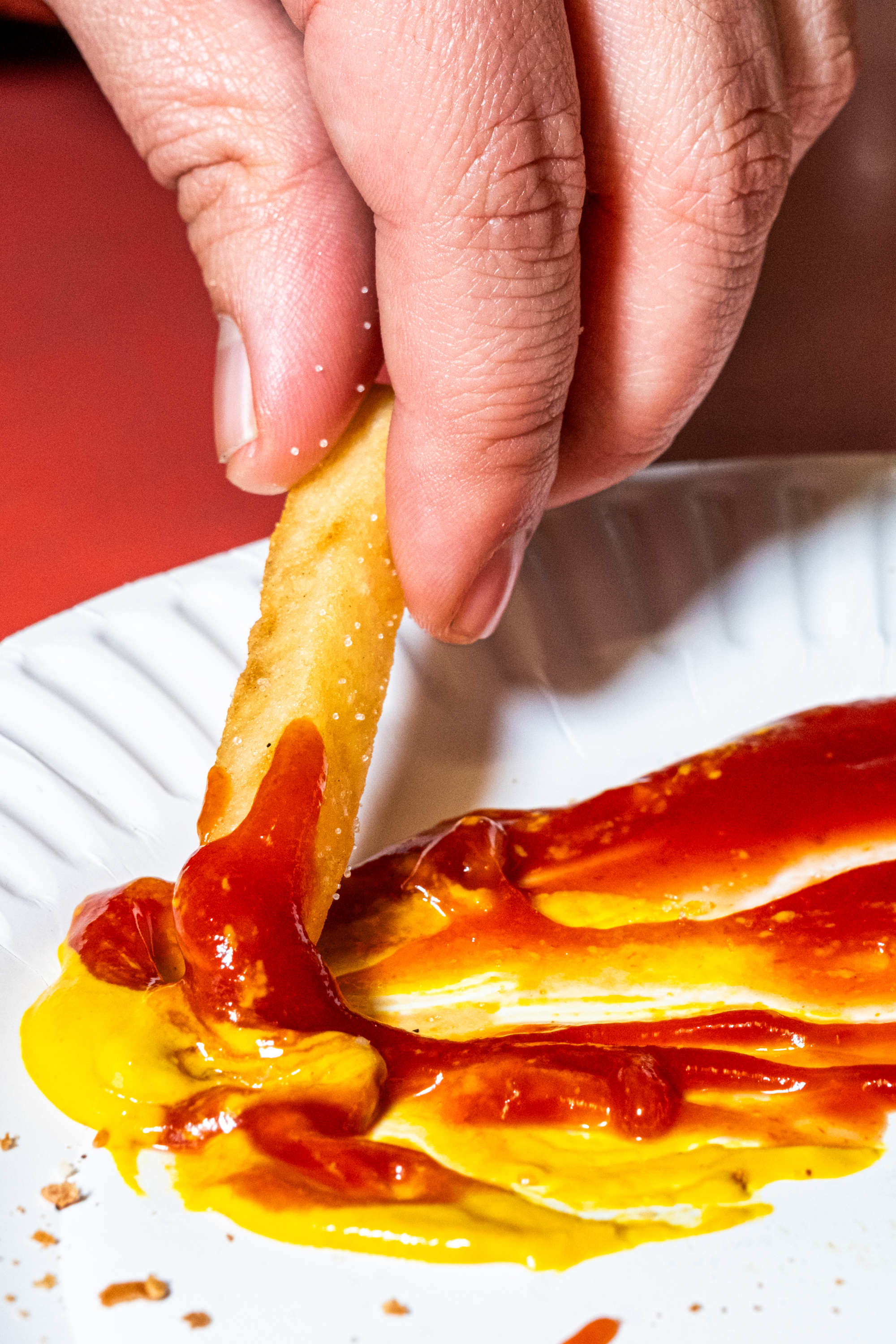 A hand holds a French fry, dipping it into a mix of ketchup and mustard on a white paper plate, with crumbs scattered nearby.