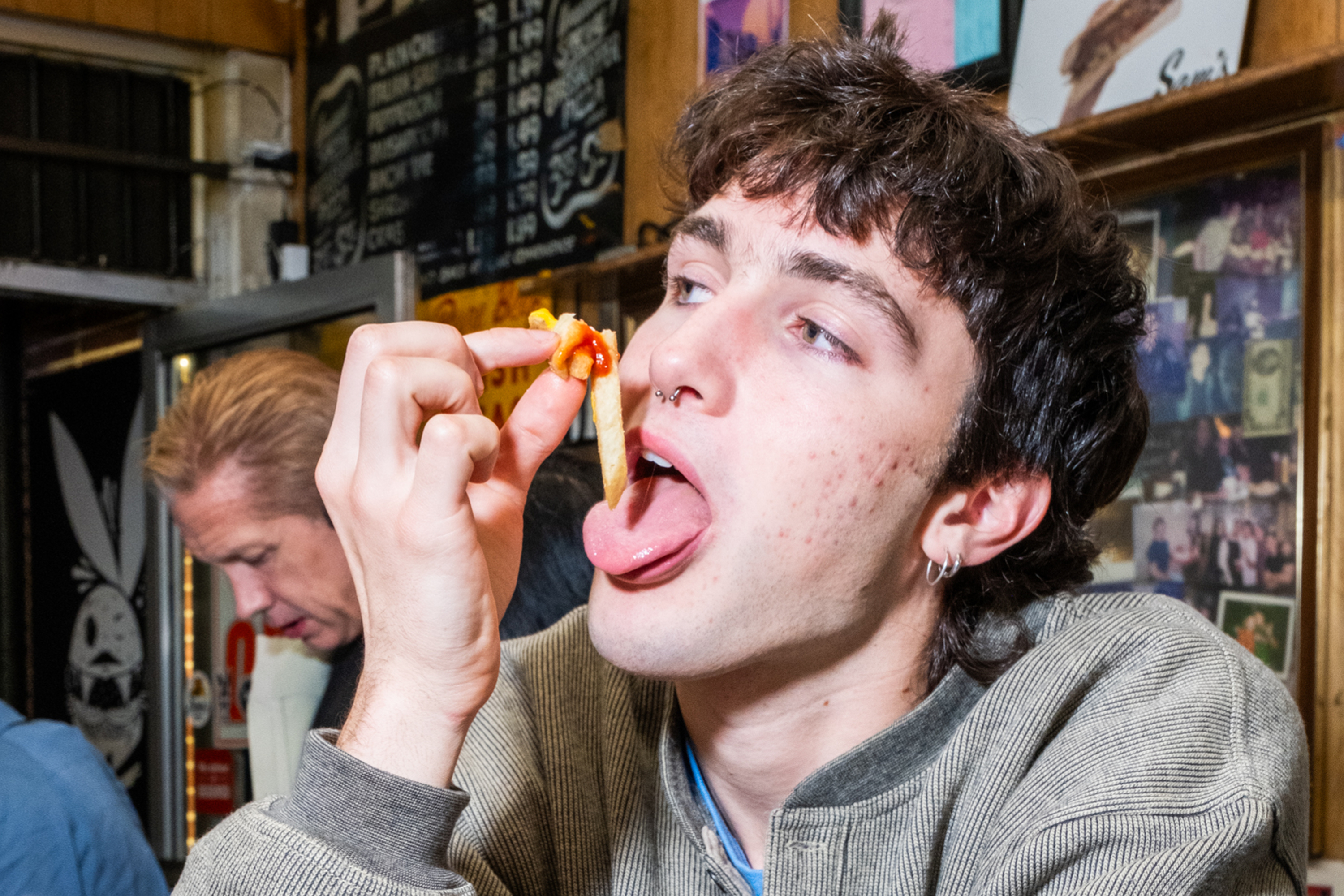 A person with short hair and earrings is holding a french fry with tomato sauce near their mouth in a diner, with a man blurred in the background.