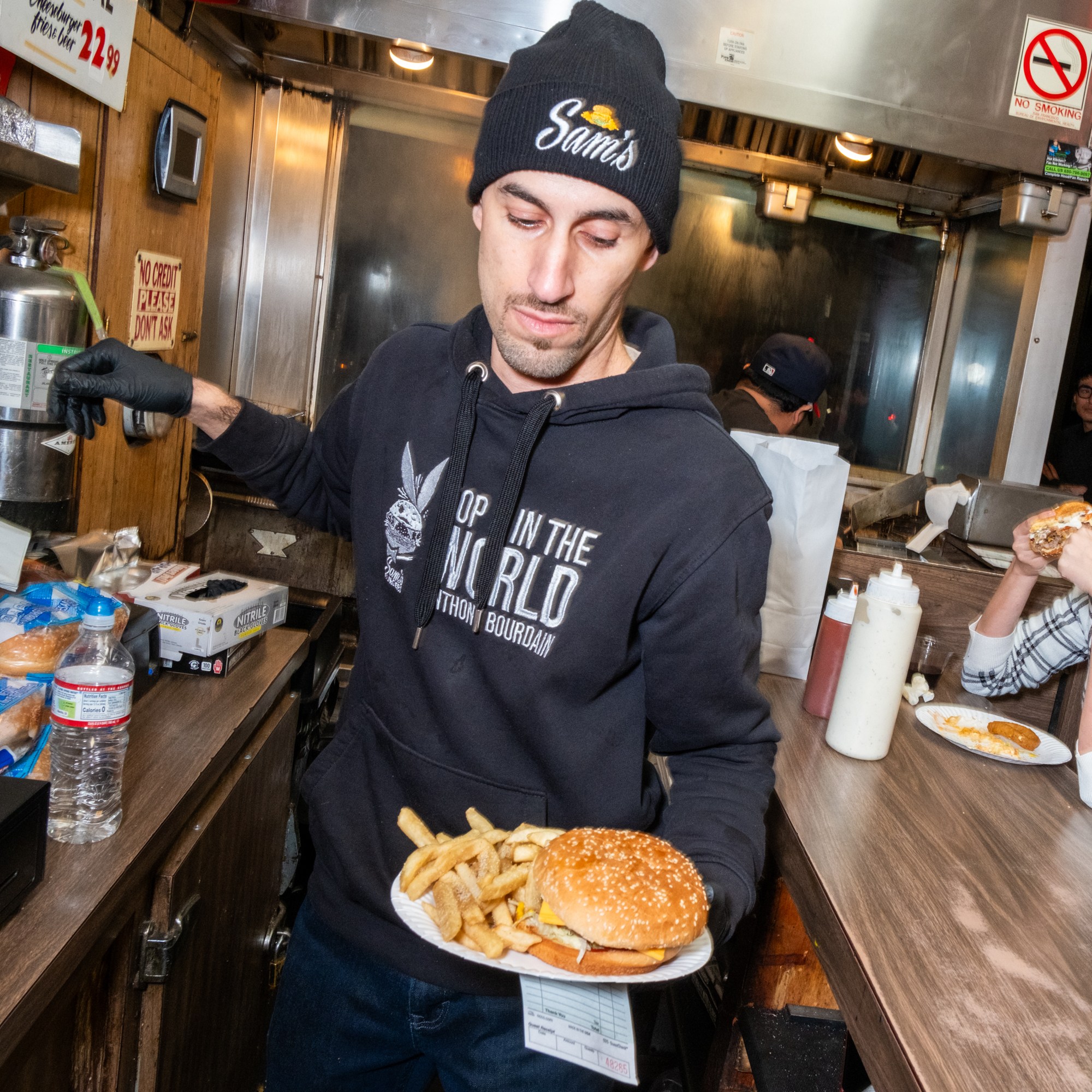 A man in a beanie serves a burger and fries in a casual eatery, while a woman in a plaid shirt sits at the counter eating her meal. Signs and condiments are visible.