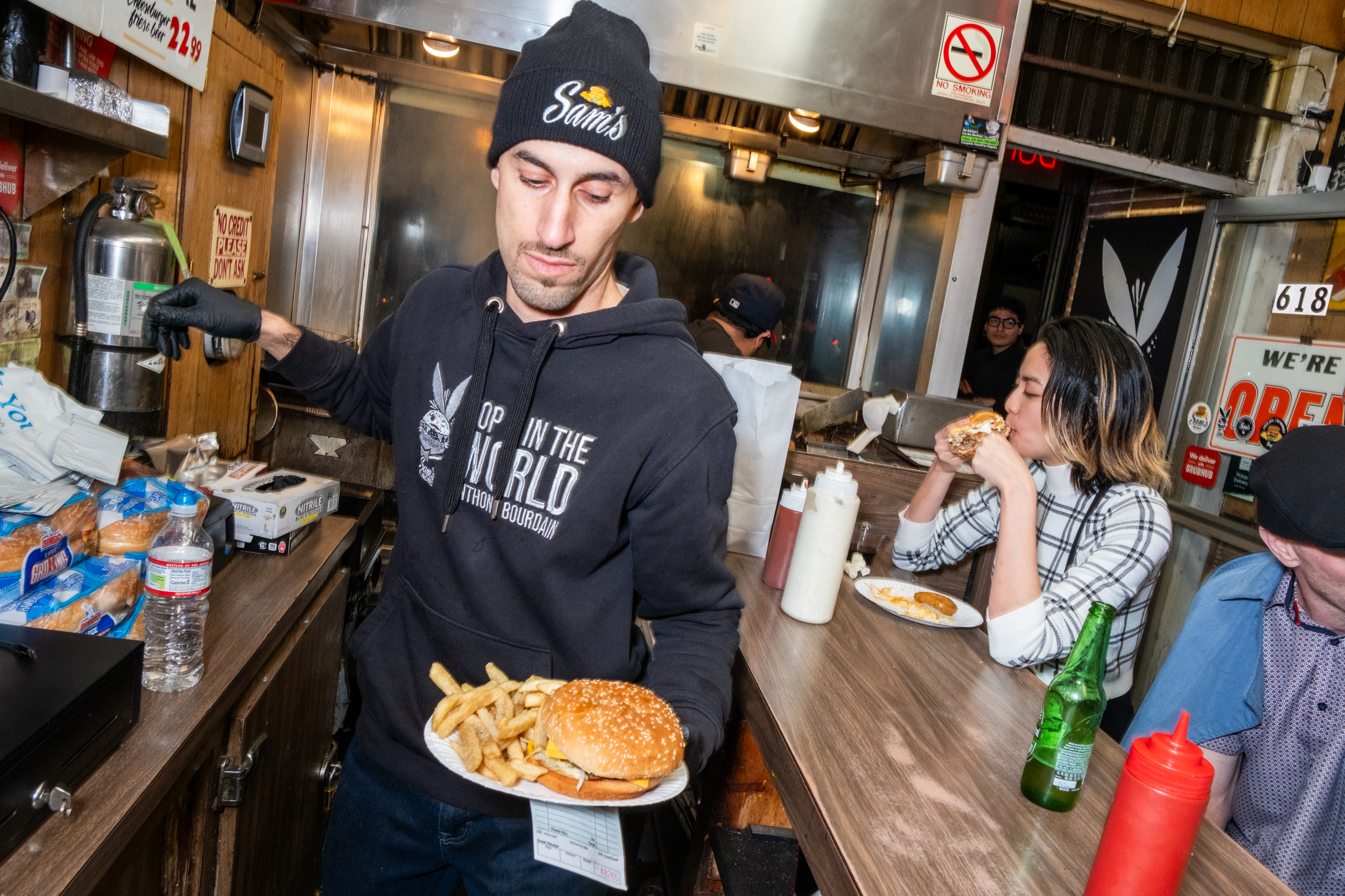 A man in a beanie serves a burger and fries in a casual eatery, while a woman in a plaid shirt sits at the counter eating her meal. Signs and condiments are visible.