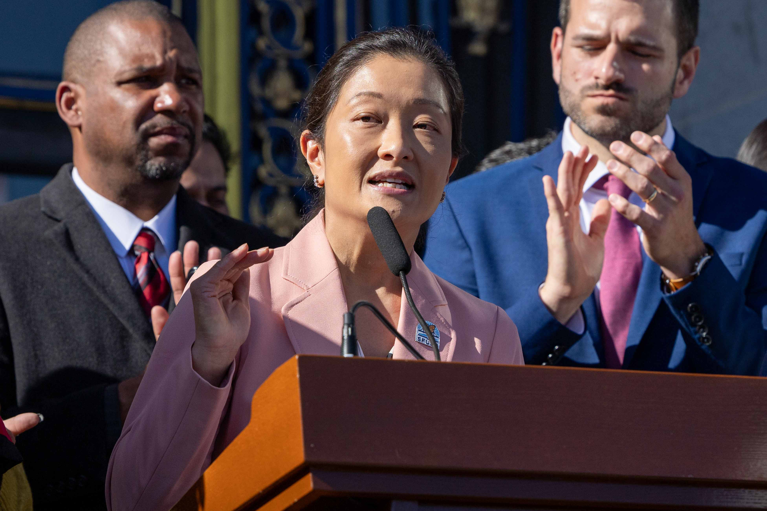 A woman in a pink blazer speaks at a podium, flanked by two men clapping. The setting appears formal, possibly a public announcement or speech.