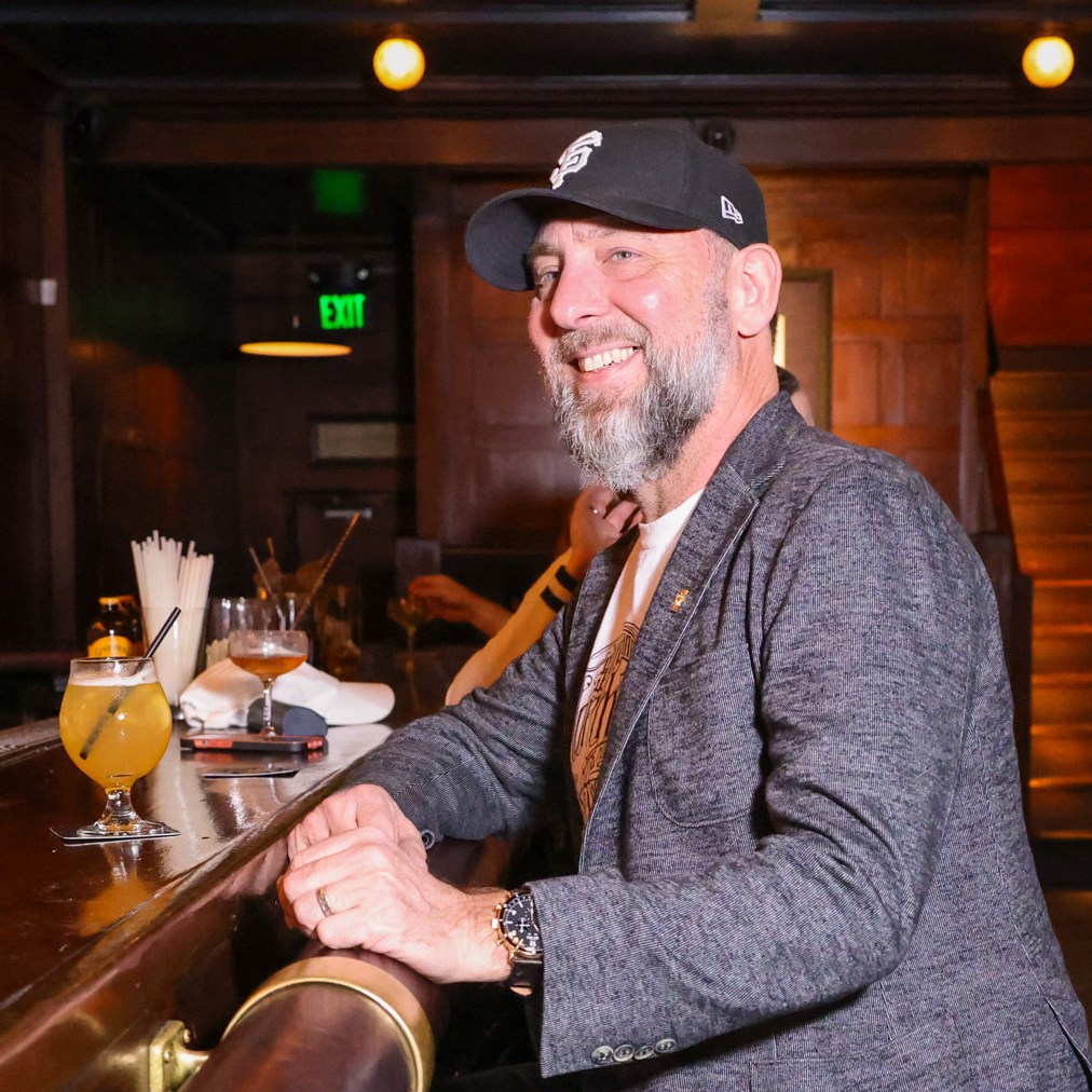 A smiling man with a beard and a cap is sitting at a bar with a drink in front of him. The background features wooden paneling and a staircase.