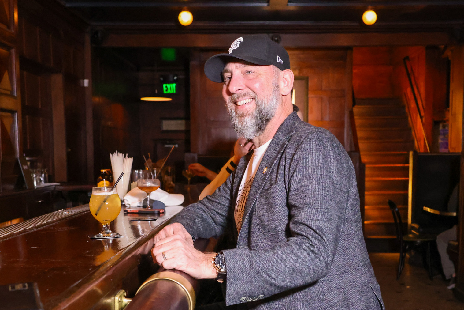 A smiling man with a beard and a cap is sitting at a bar with a drink in front of him. The background features wooden paneling and a staircase.