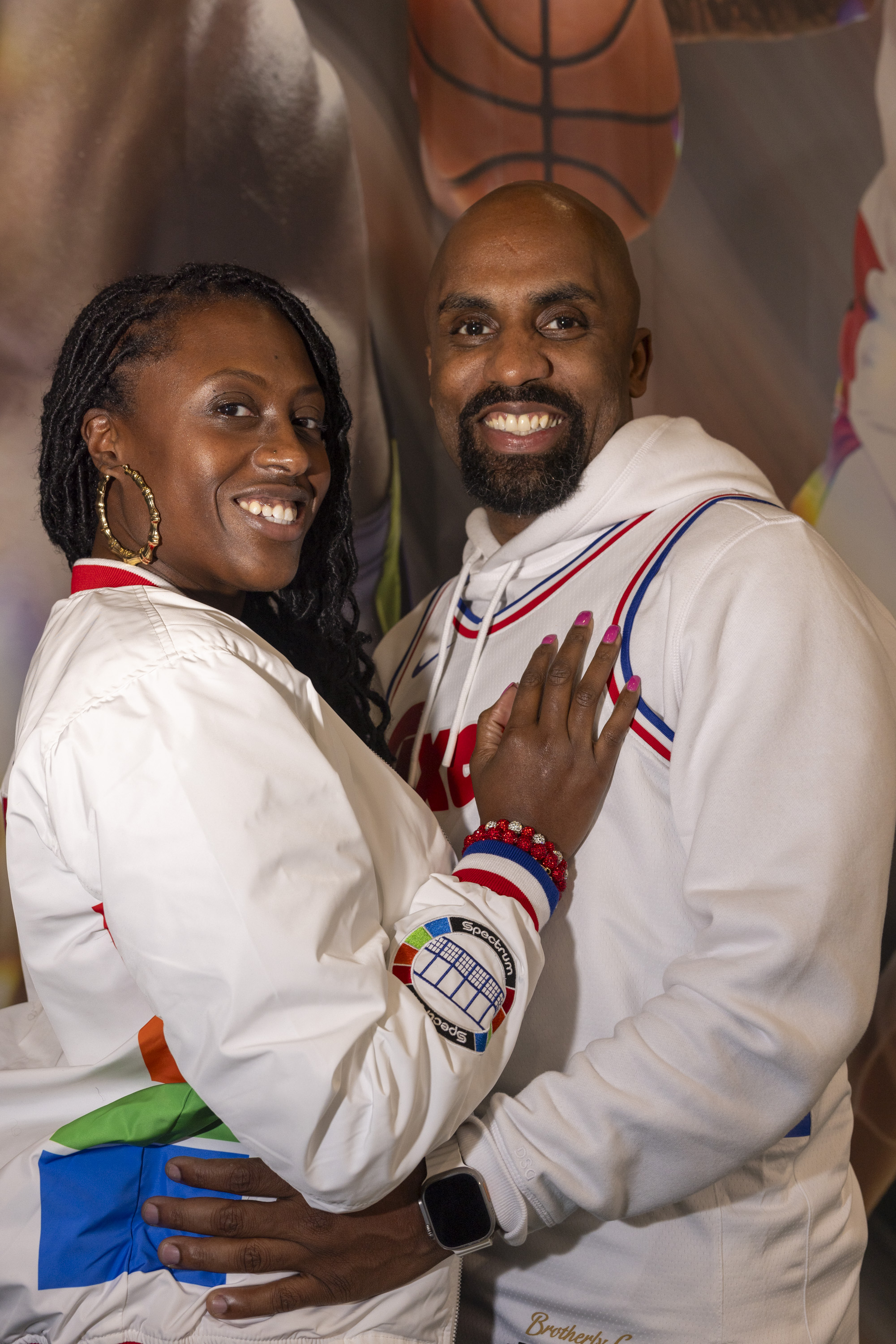 A smiling couple poses together in matching white outfits. The woman has long hair and wears hoop earrings. A basketball image is seen in the background.