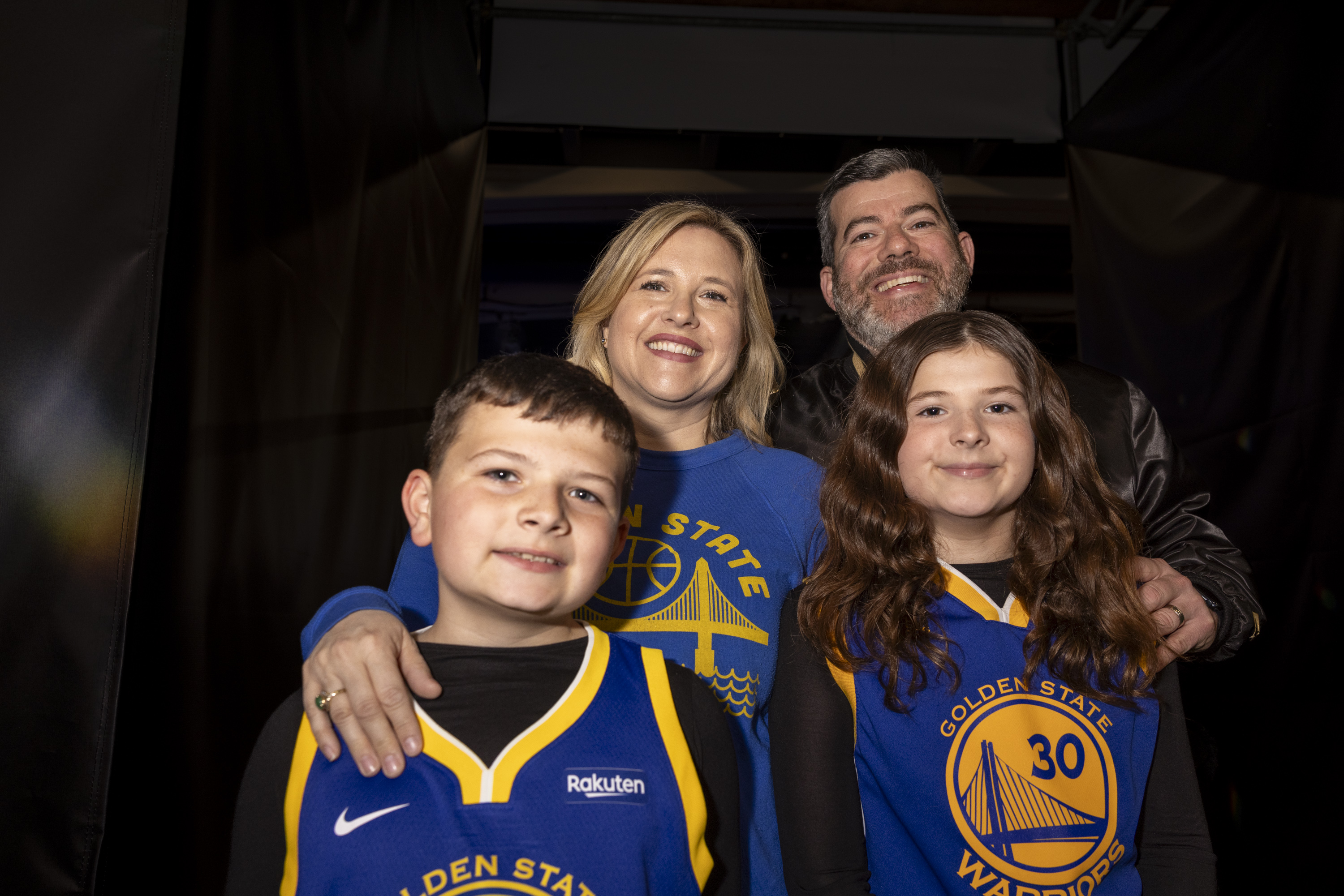 A smiling family of four is pictured wearing Golden State Warriors shirts and jerseys. They are standing closely together in a warmly lit setting.