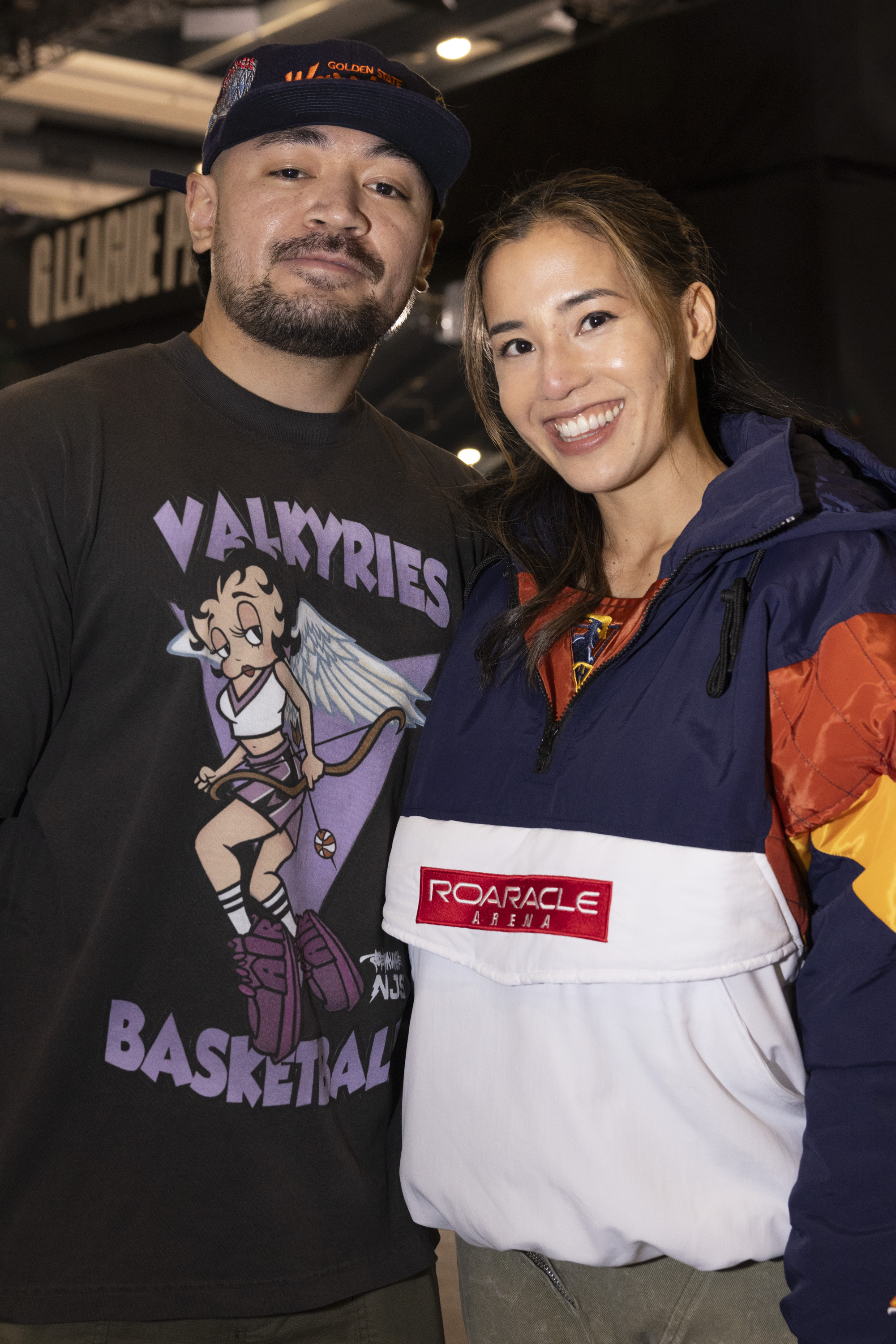 A smiling man and woman are posing. The man wears a cap and a t-shirt featuring a cartoon character and &quot;Valkyries Basketball.&quot; The woman wears a jacket with &quot;ROARacle Arena.&quot;