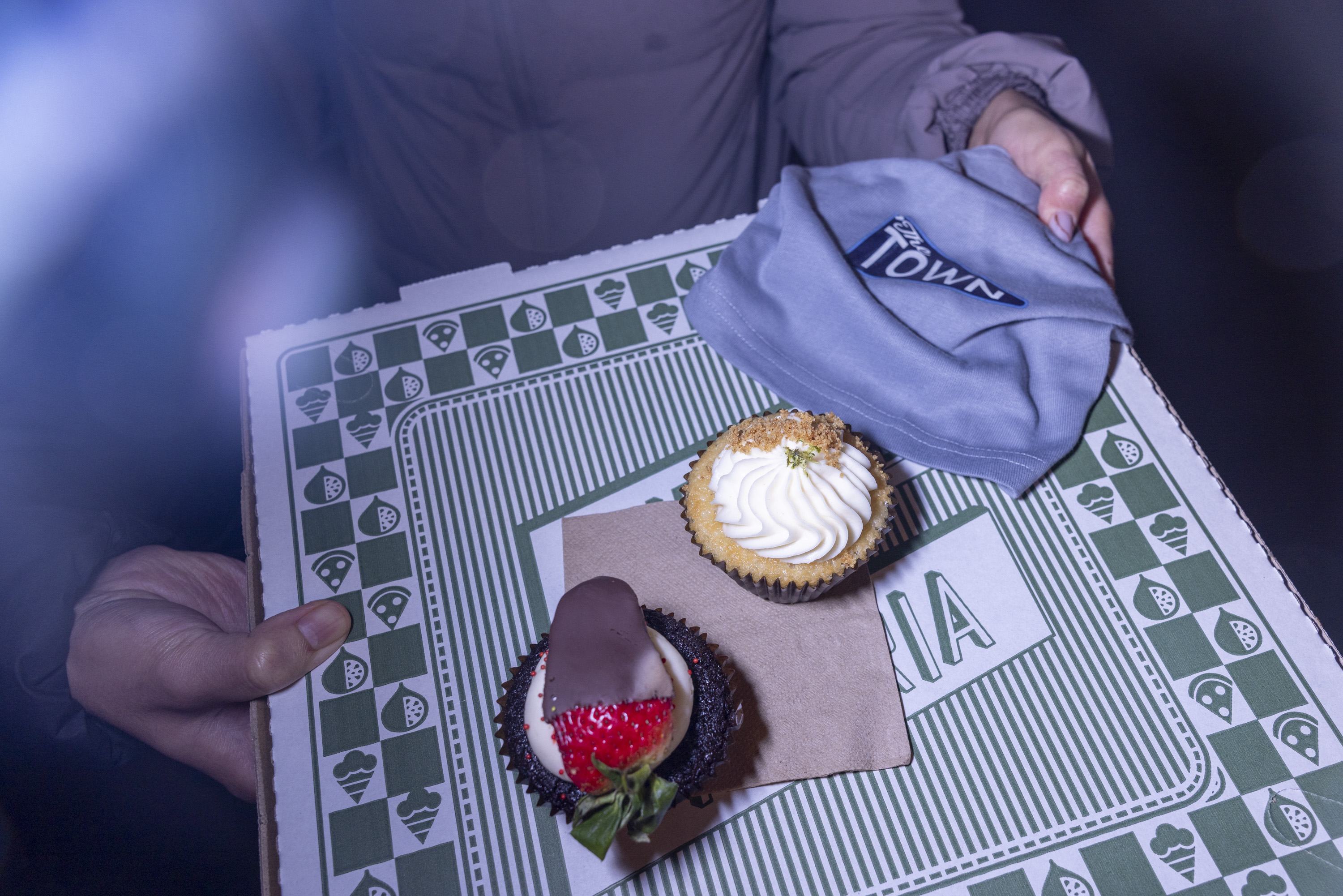 Two cupcakes sit on a pizza box; one topped with chocolate and a strawberry, the other with cream and crumbs. A gray cloth with &quot;Town&quot; is nearby.