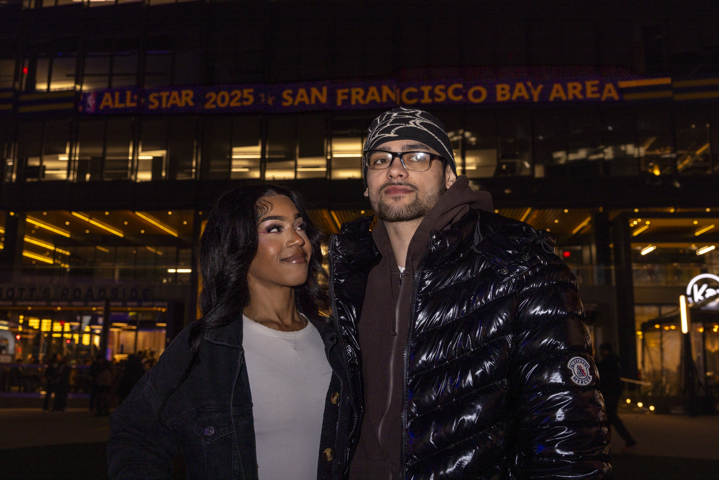 A couple stands outside at night in front of a lit building with a sign reading &quot;All-Star 2025 San Francisco Bay Area.&quot; The woman smiles at the man.