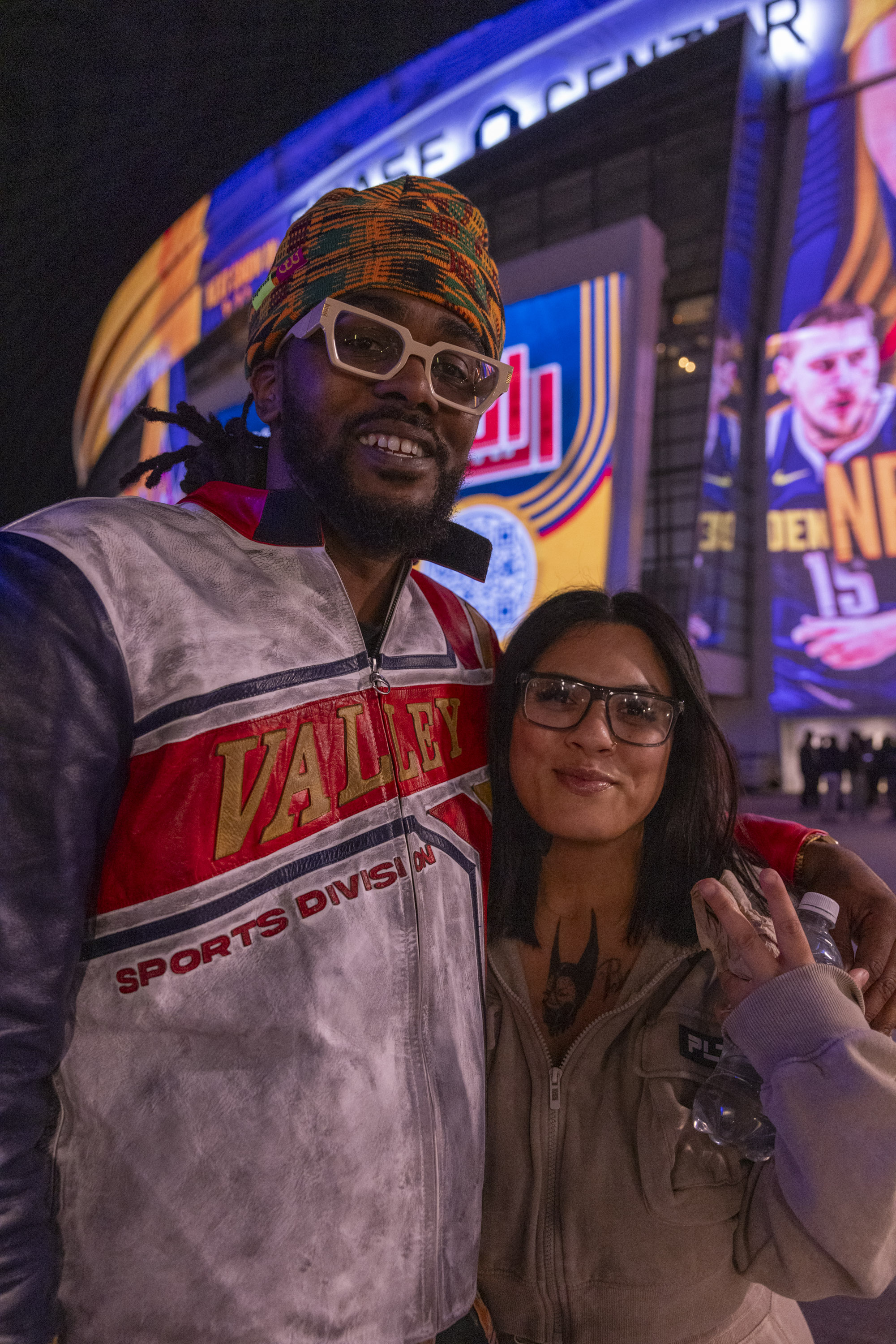 A smiling man and woman pose for a photo outside an arena displaying bright sports graphics. The man wears a colorful headwrap and jacket, the woman a hoodie.