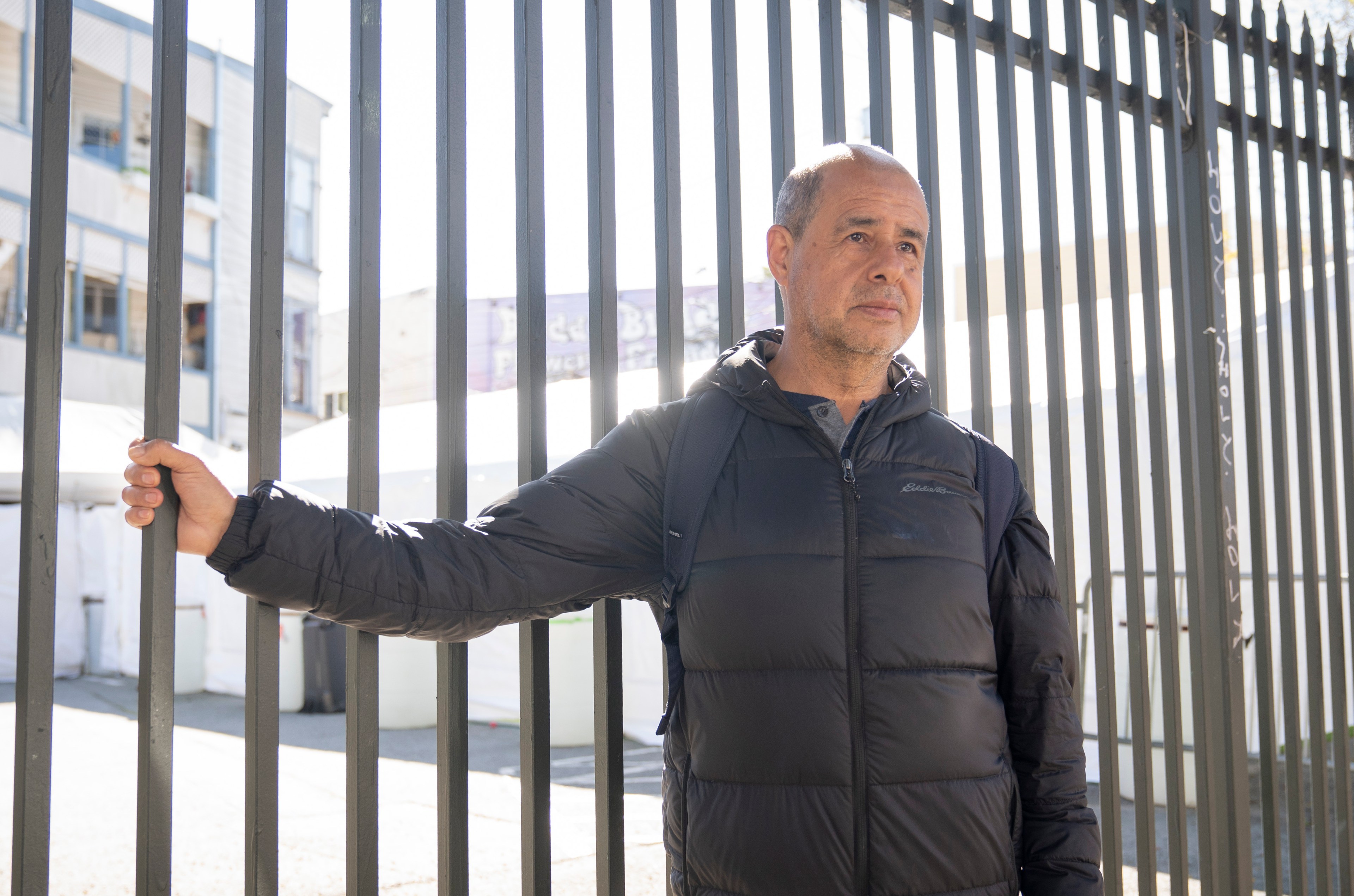 A man in a black puffer jacket stands next to a tall metal fence, holding it with one hand. The background shows a street and buildings in daylight.