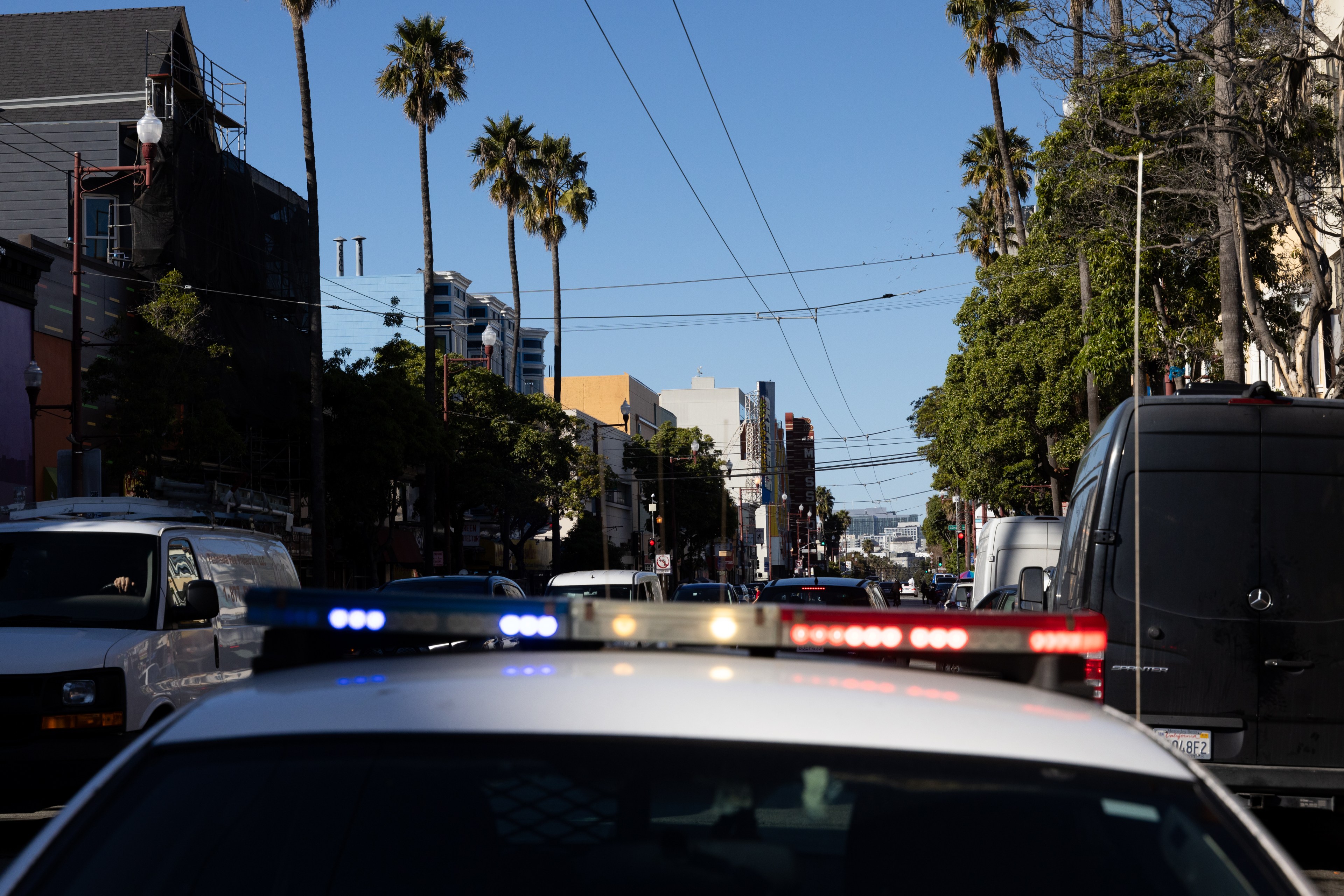 A street scene with tall palm trees, various buildings, and a mix of vehicles. A police car with red and blue lights is in the foreground.