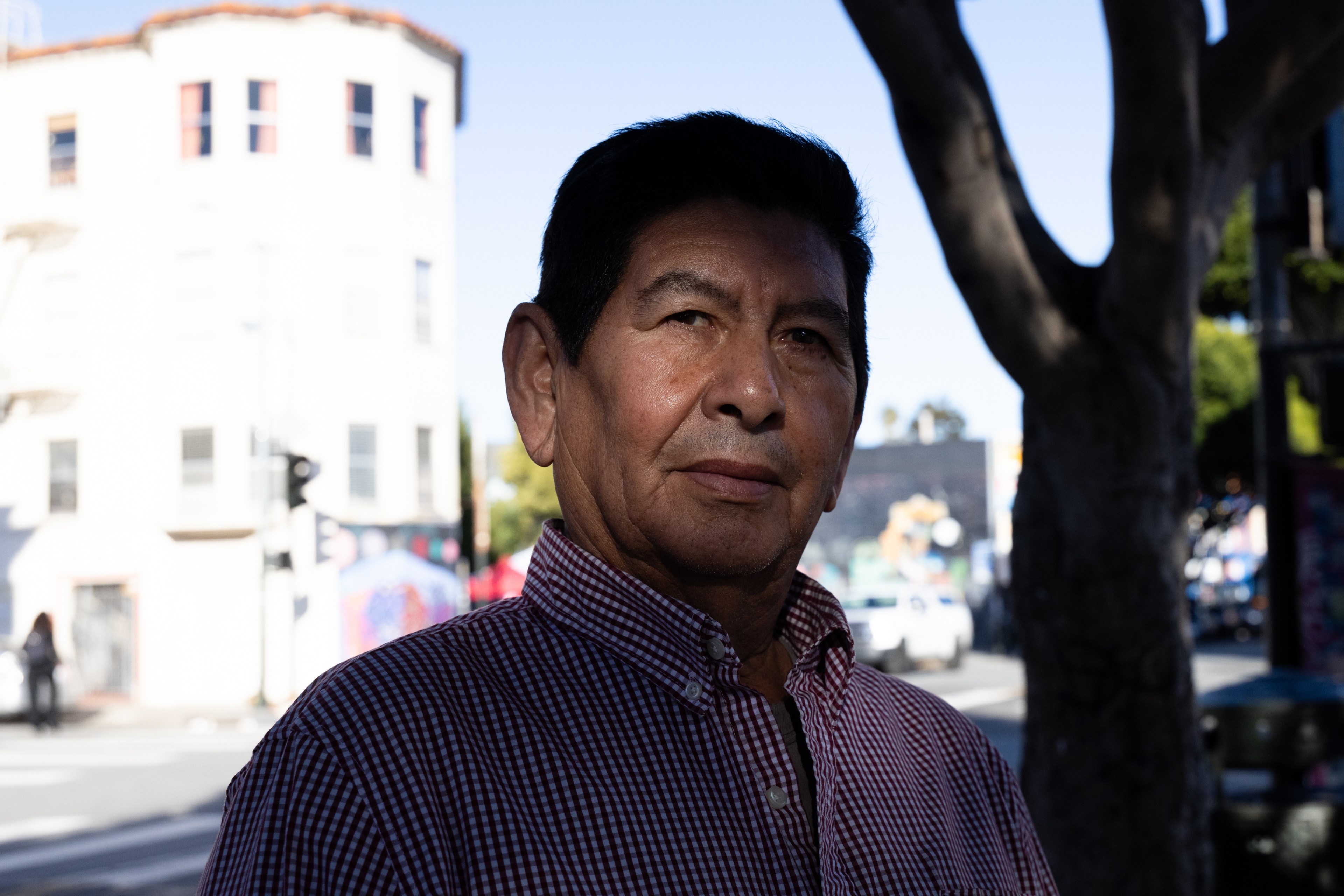 A man in a checkered shirt stands on a city street near a tree. The background features a brightly lit white building and blurred street elements.