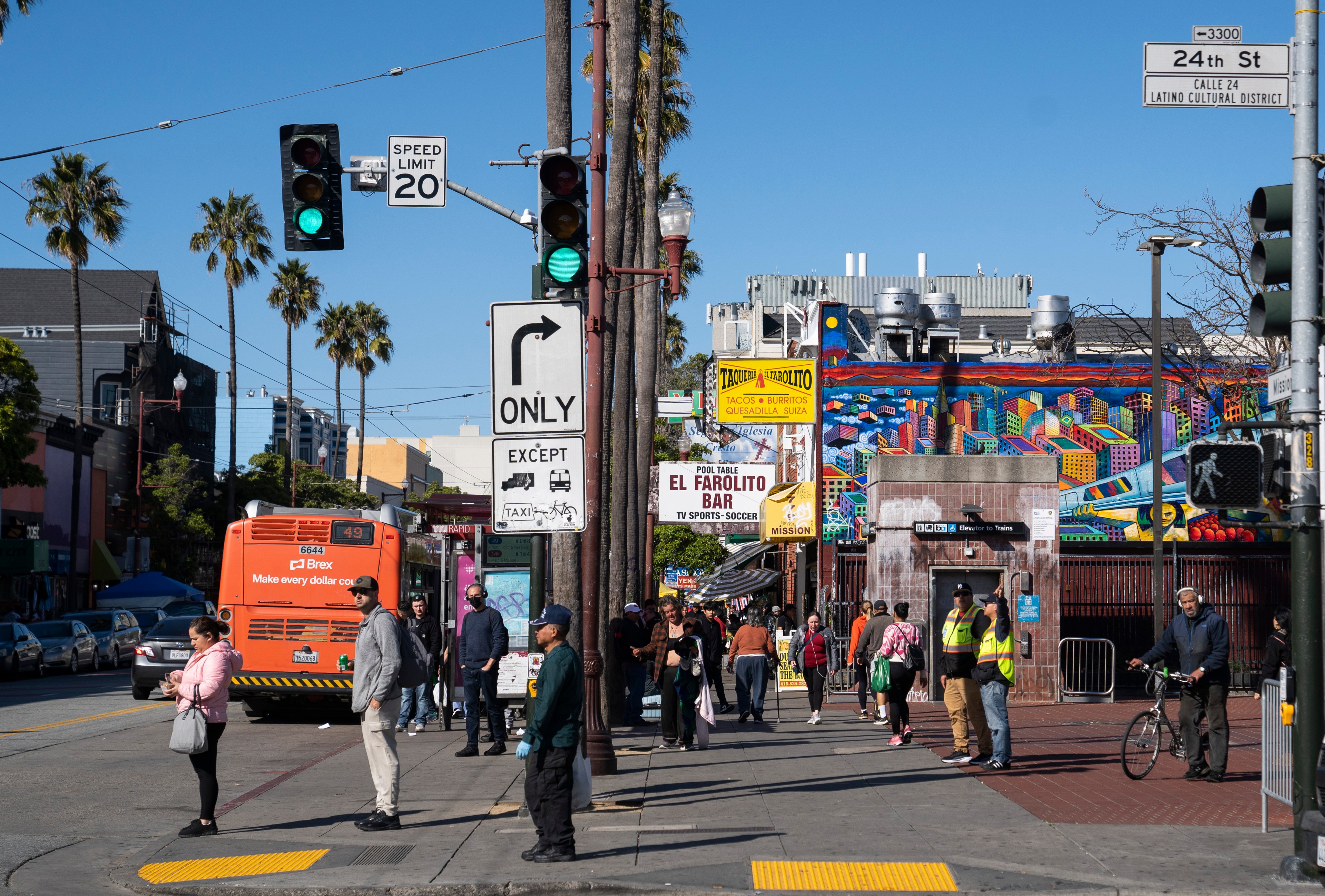 A busy street corner with people walking, a bus, palm trees, and vibrant murals. Signs indicate it's the Latin Cultural District on 24th Street.