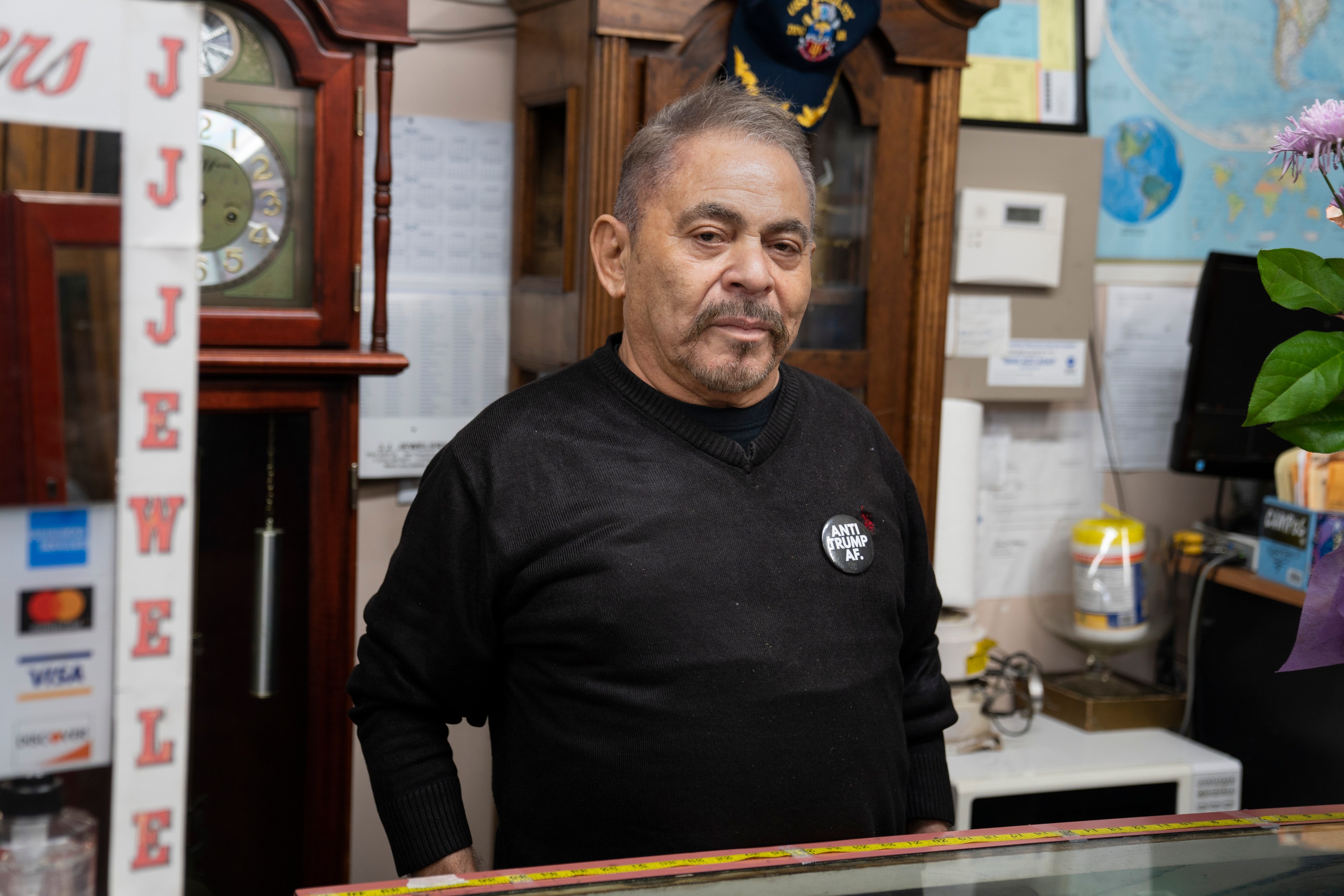 A man in a black sweater stands behind a counter with a &quot;JEWELERS&quot; sign and wears a badge that says &quot;ANTI TRUMP AF.&quot; A clock and world map are visible behind him.