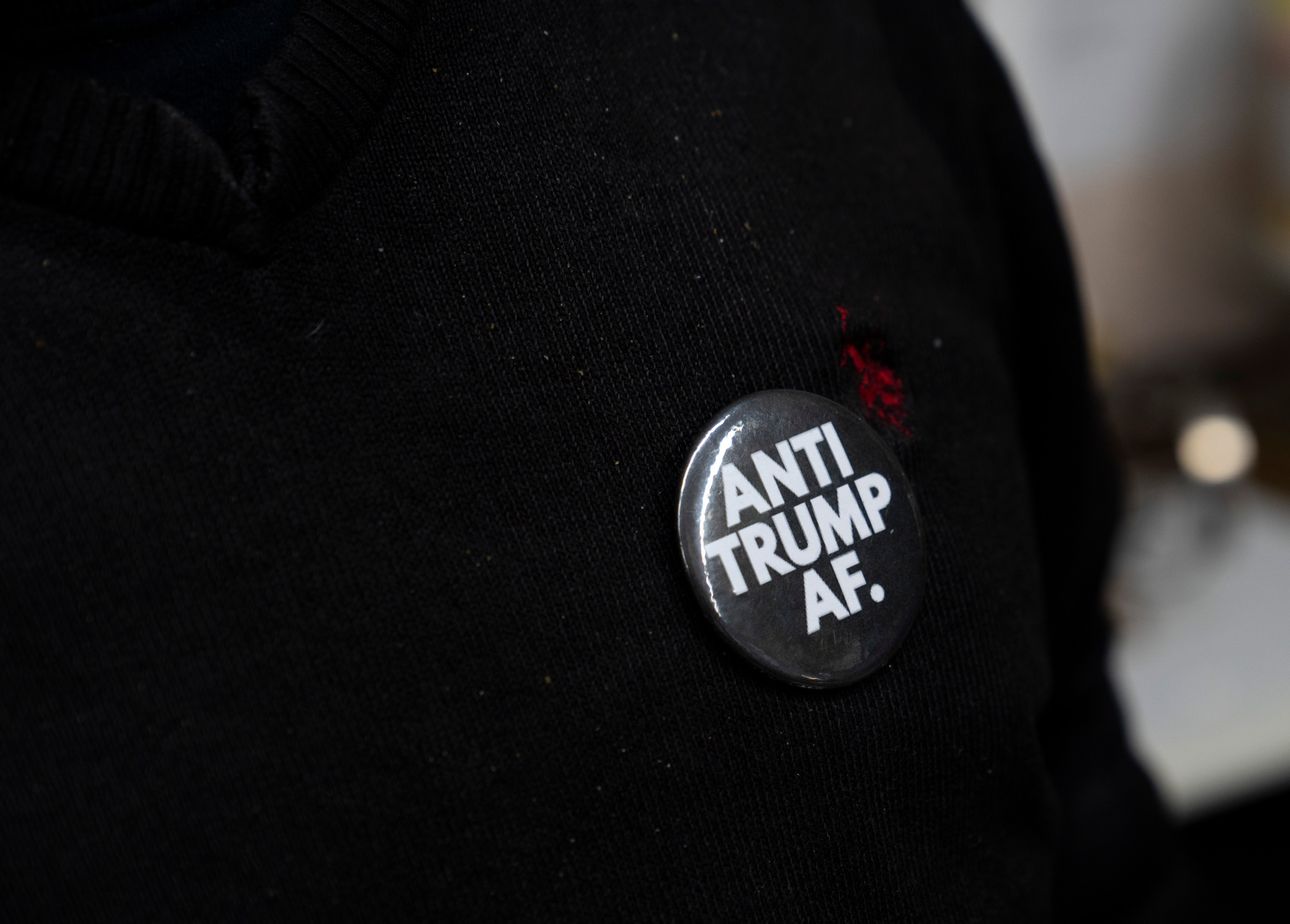 A person wearing a black shirt has a button pinned to it that reads &quot;ANTI TRUMP AF&quot; in bold white letters.