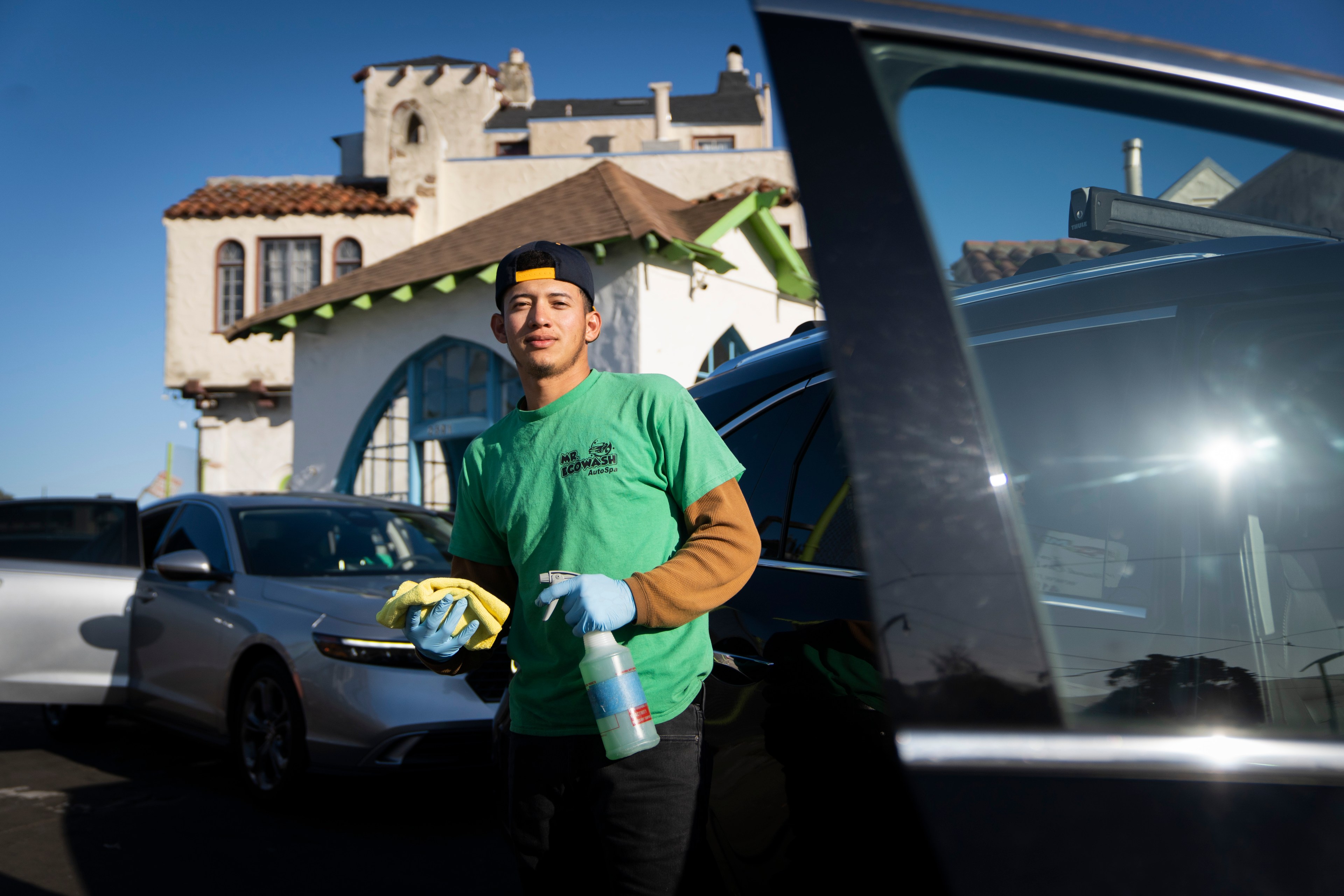 A person is standing beside a car, wearing a green shirt and gloves, holding cleaning supplies. A building with a sloped roof is in the background.