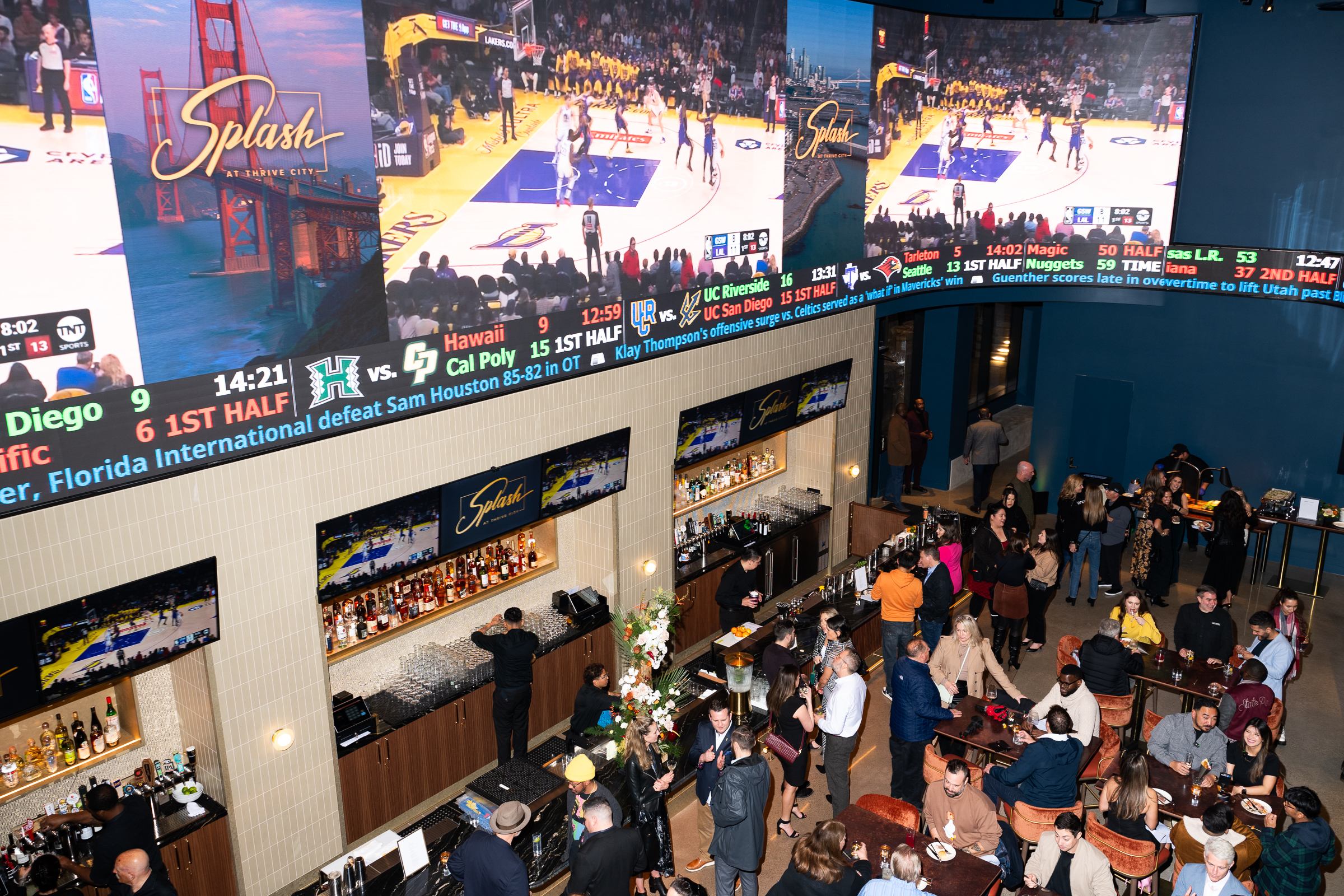 A lively bar scene with people socializing and watching sports on large screens displaying basketball and other games. The bar is named "Splash" at Thrive City.