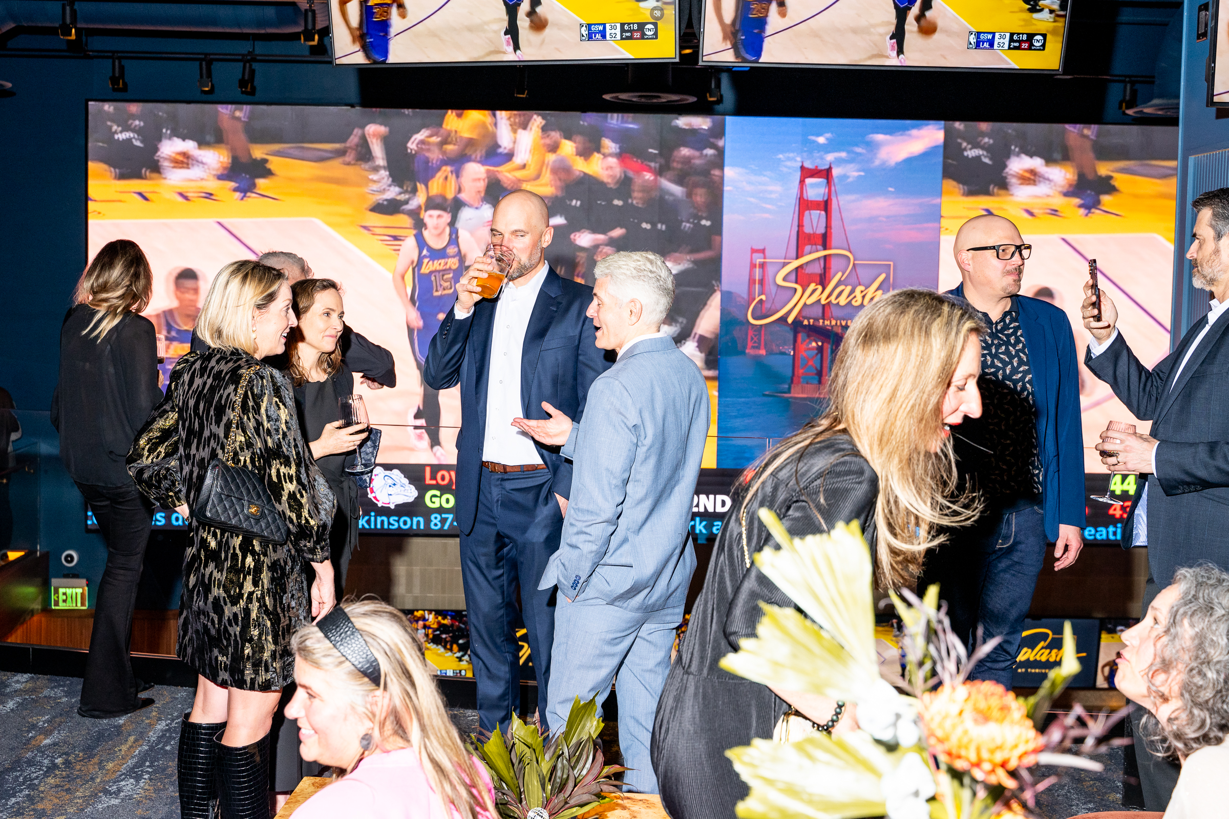 People are socializing at a lively event with large screens displaying sports and scenic graphics. Some hold drinks while engaging in conversation.