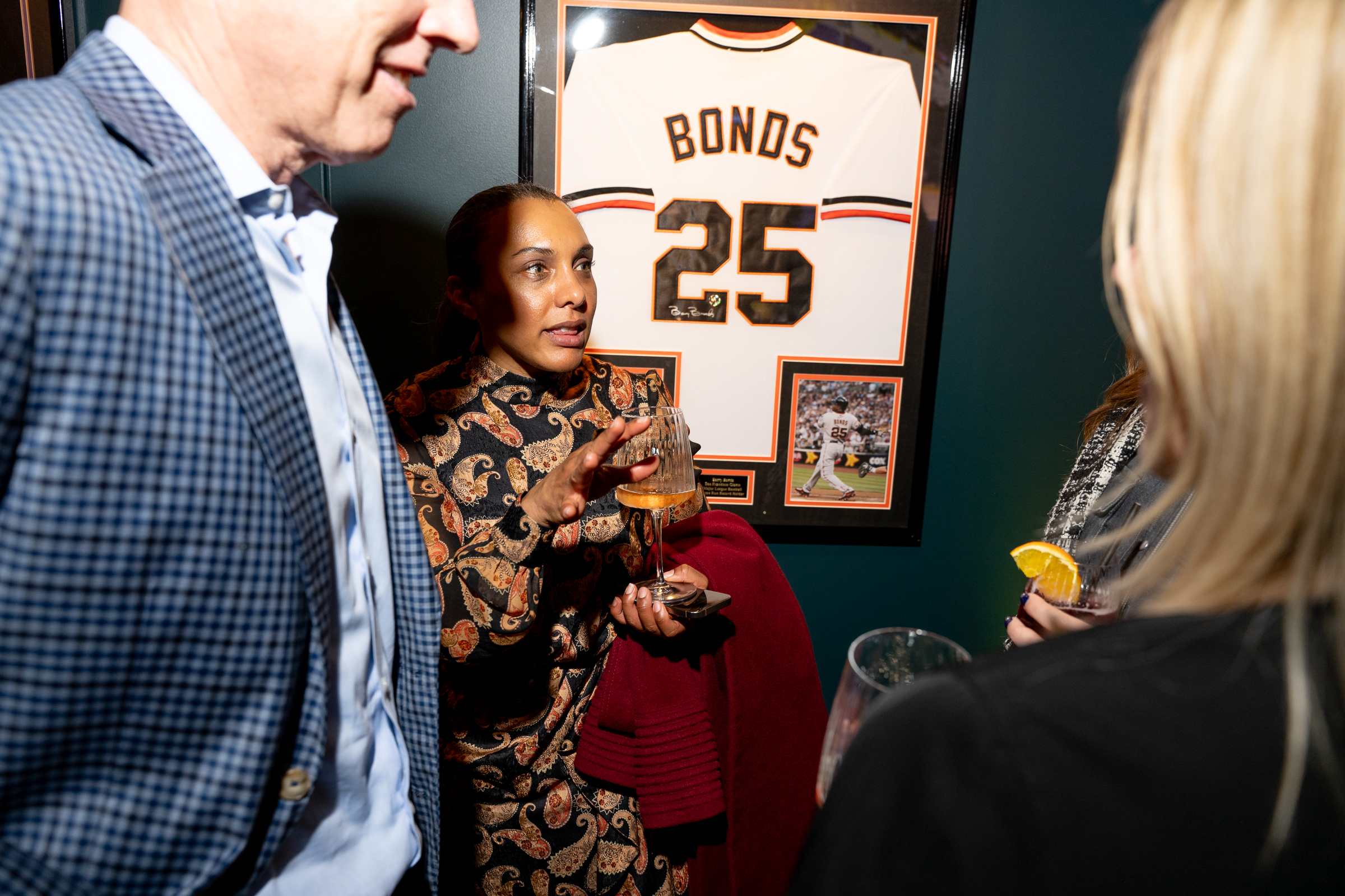 Three people are engaged in conversation, holding drinks. Behind them is a framed sports jersey with the name &quot;Bonds&quot; and the number 25 on it.