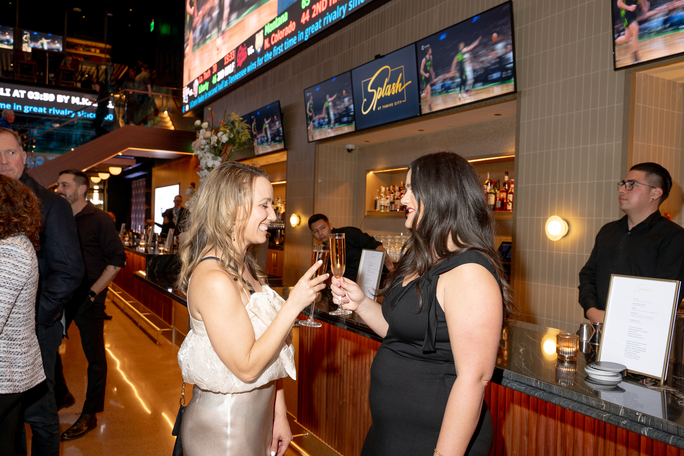 Two women are toasting with champagne glasses in a lively bar. Behind them, a bartender stands near bottles, and TV screens display sports images.