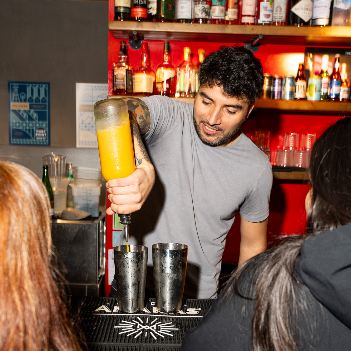 A bartender with a tattoo pours orange liquid into a shaker at a bar. Behind him, shelves hold various bottles. Two people sit at the counter.