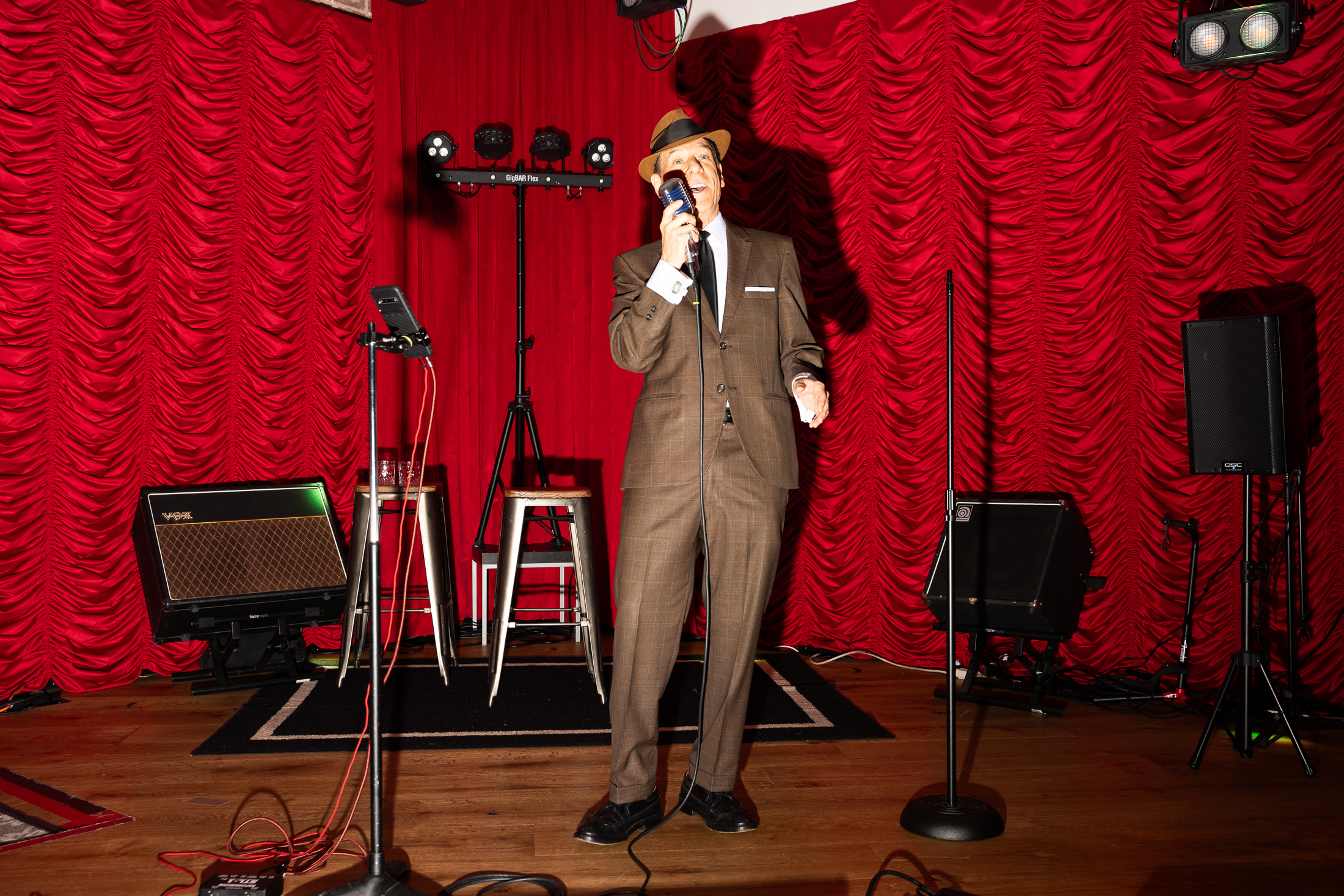 A person in a brown suit and hat stands on a stage with red curtains, holding a microphone. The stage is equipped with speakers, lights, and musical equipment.