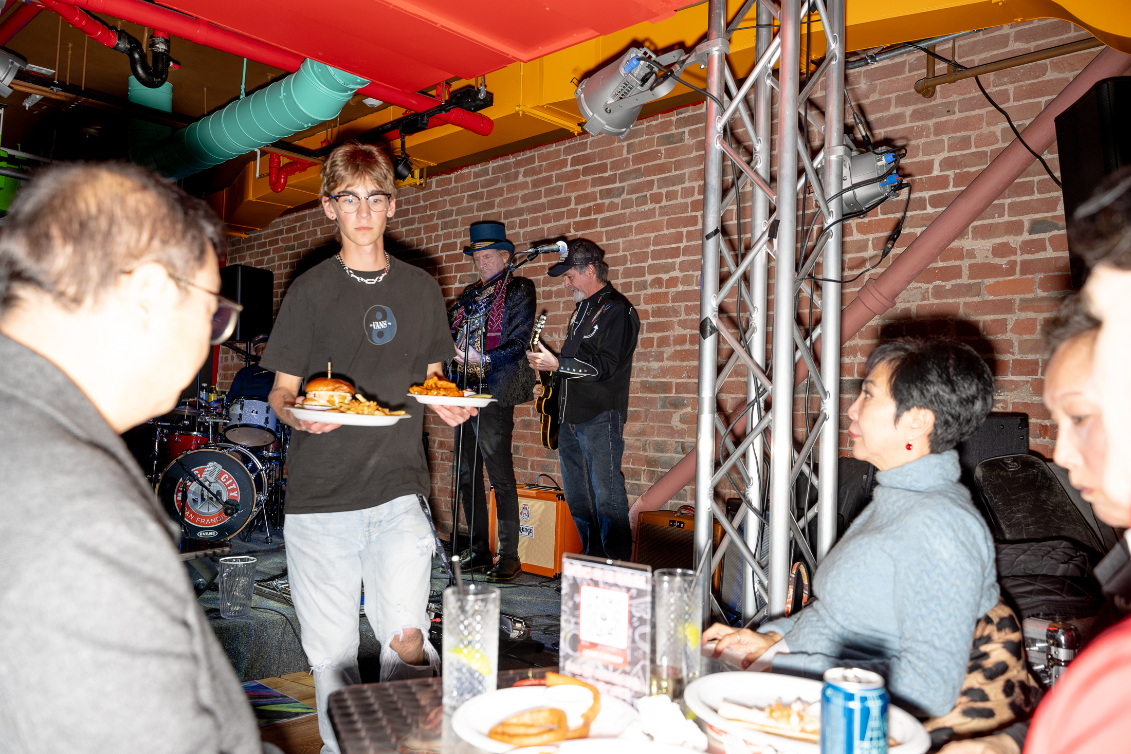 A young man carries plates of food in a vibrant restaurant with people seated at a table in the foreground, while musicians perform on a small stage in the background.