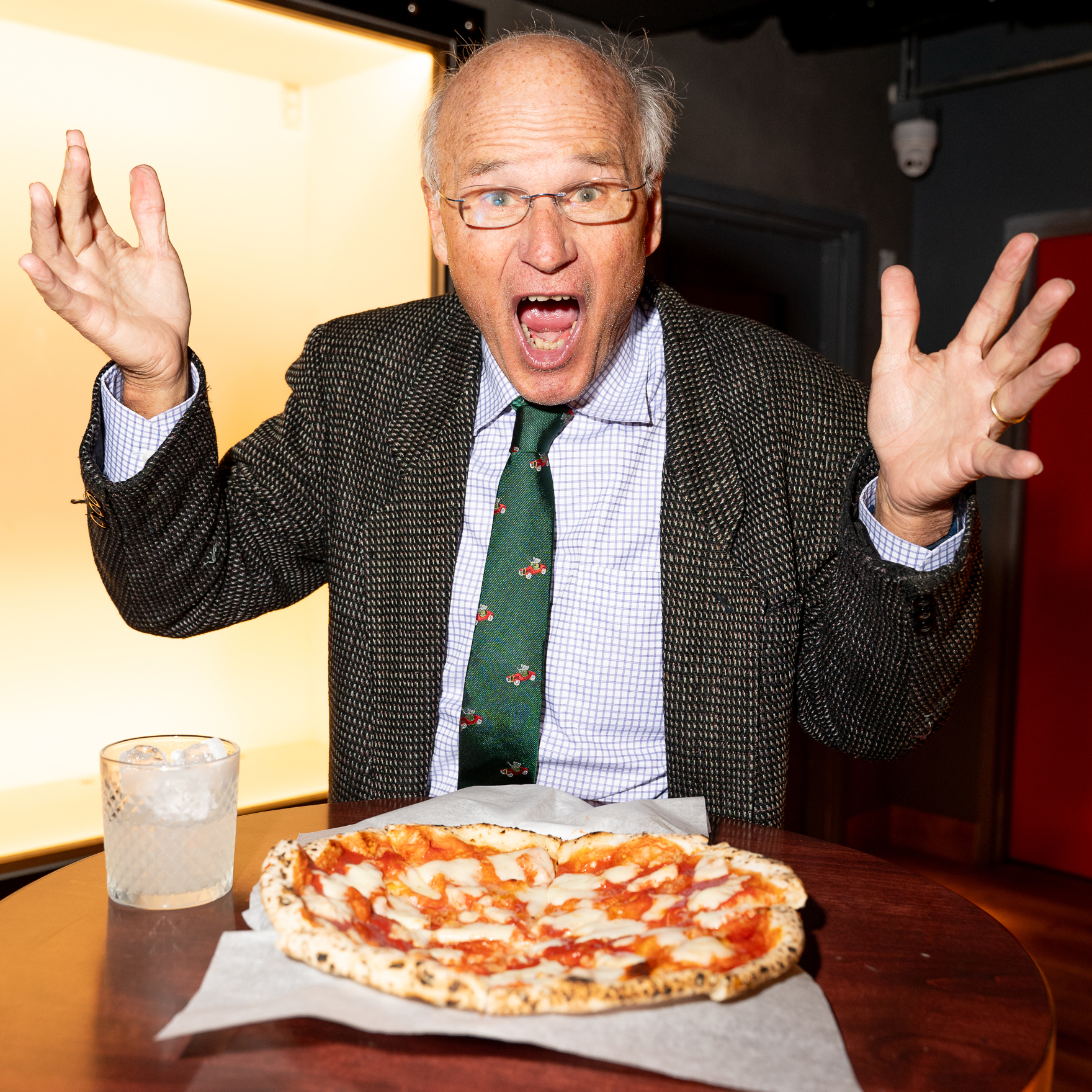 A man in a suit and tie, expressing surprise or excitement, is seated at a table with a pizza and a glass of ice water in front of him.