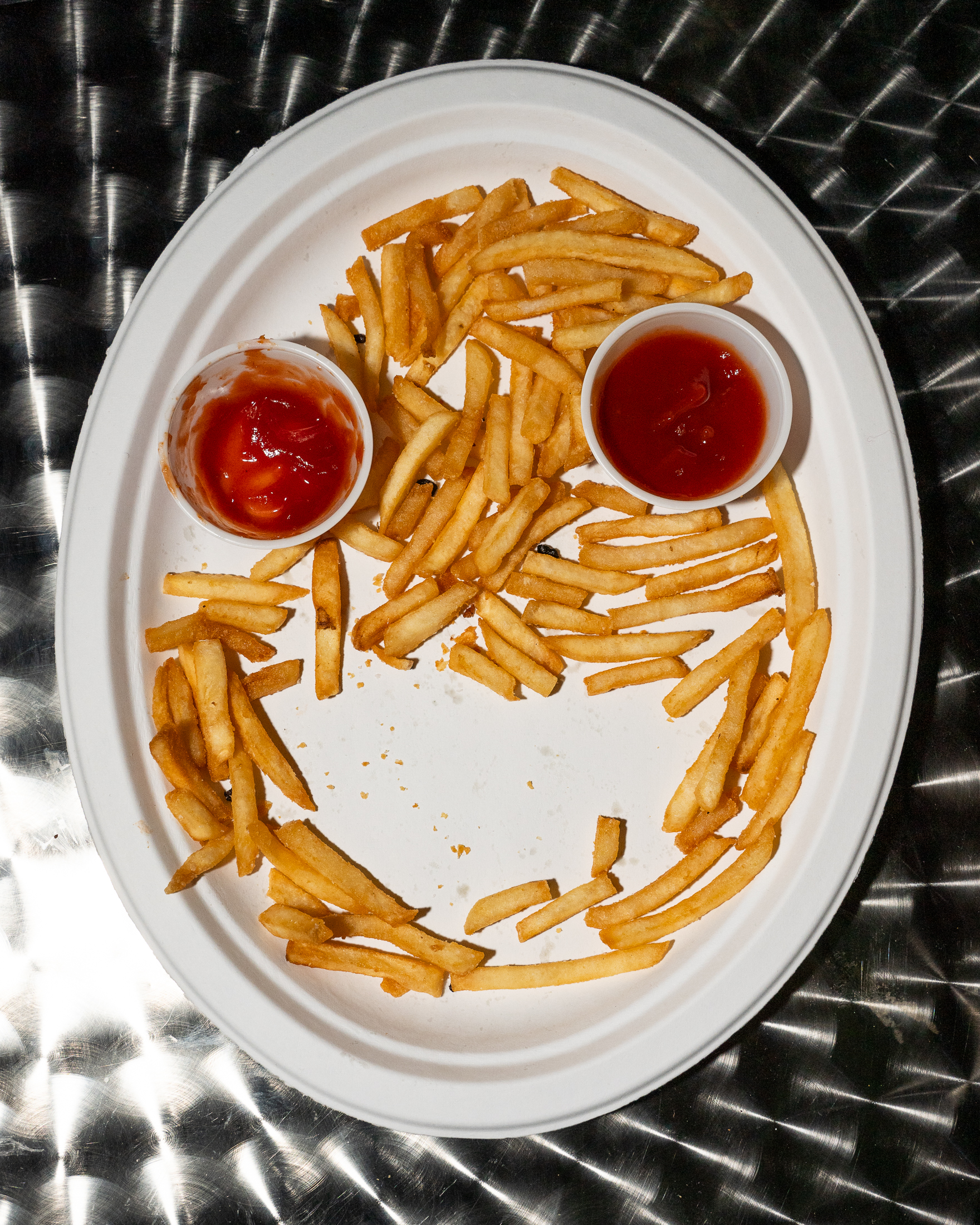 A white oval plate holds French fries arranged to form a smiley face, with two cups of ketchup as eyes. The plate is on a metallic, swirled-pattern table.