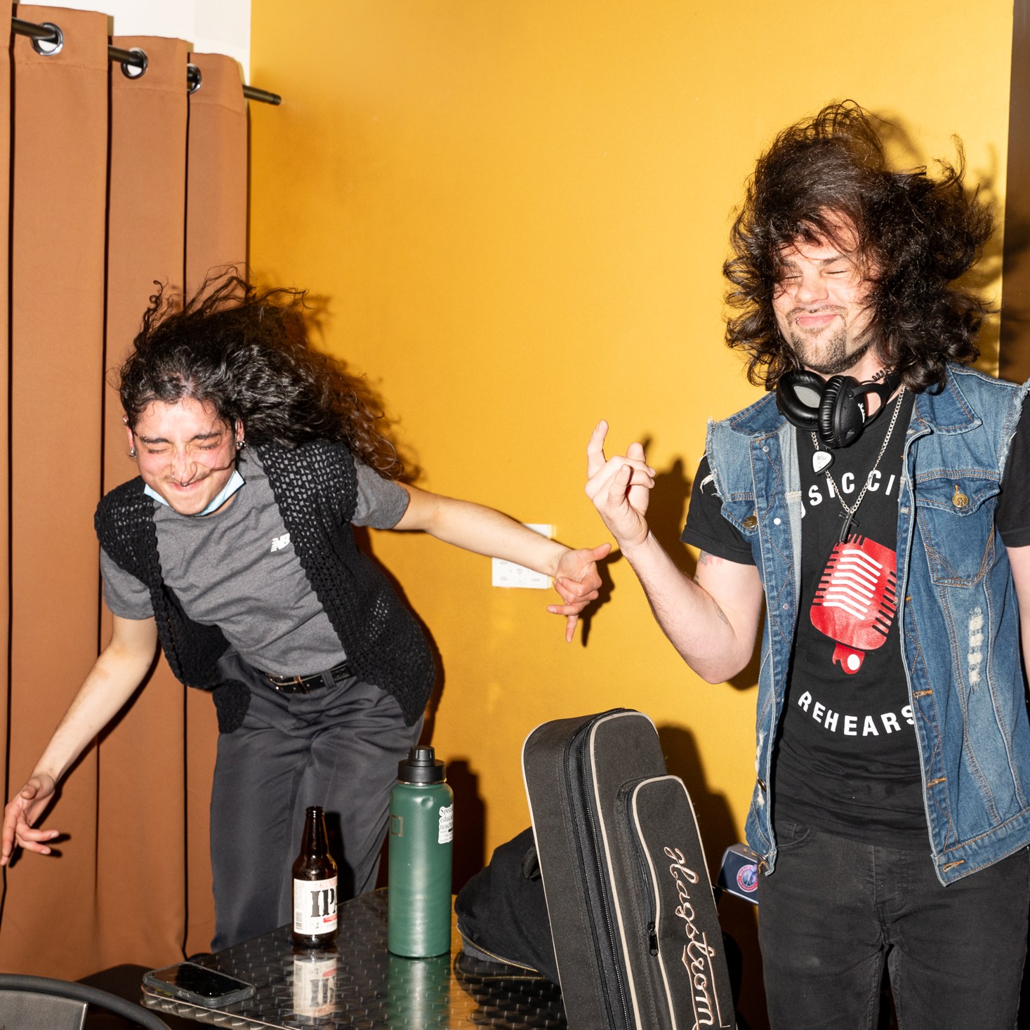 Two people are energetically dancing, hair flying, in front of a curtain. One holds a beer, the other a guitar case. A person sits in the background with a laptop.